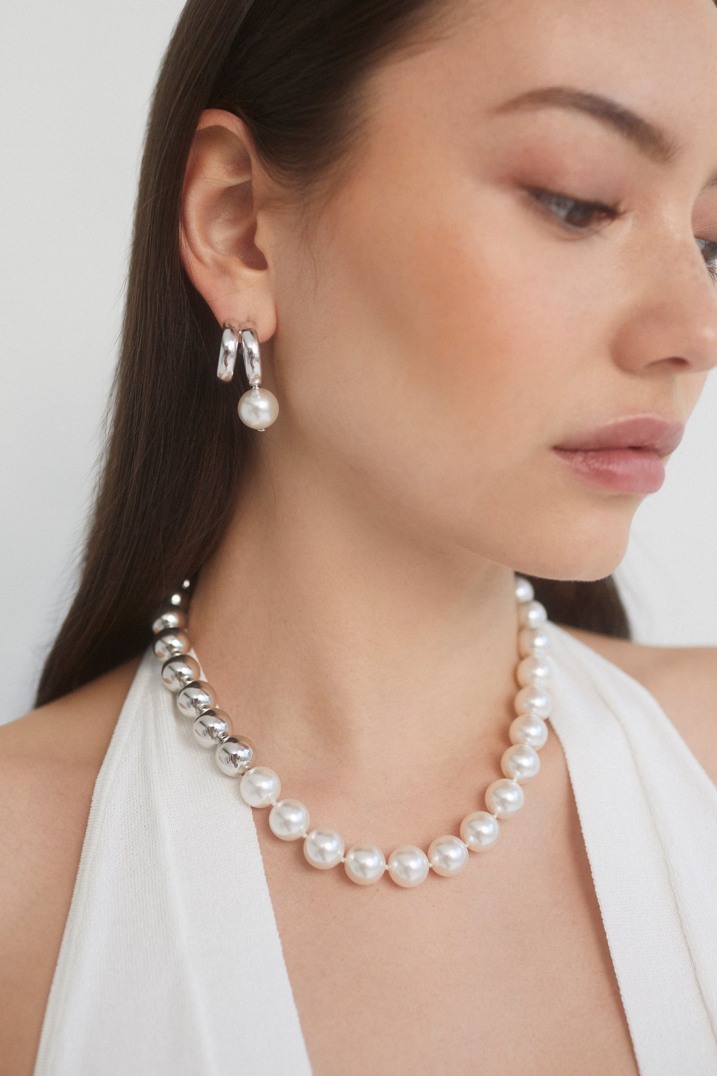 CLOSE UP IMAGE OF MODEL WEARING THE FARRAH PEARL AND METAL BEAD COLLAR NECKLACE AND FARRAH PEARL DROP DOUBLE HOOP EARRINGS IN RHODIUM PLATING WITH A WHITE HALTER TOP WHILE LOOKING OFF TO THE SIDE.