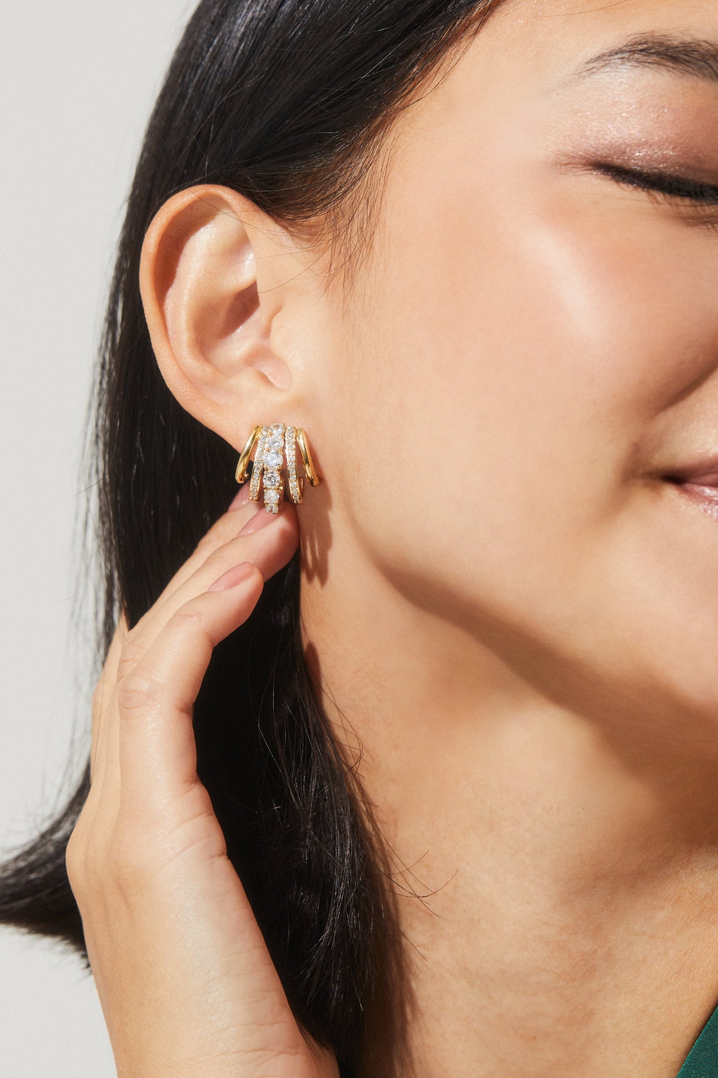 CLOSE UP IMAGE OF A MODEL WEARING THE CLEO J HOOP CZ HUGGIE EARRINGS IN GOLD PLATING WHILE GENTLY TOUCHING HER EAR.