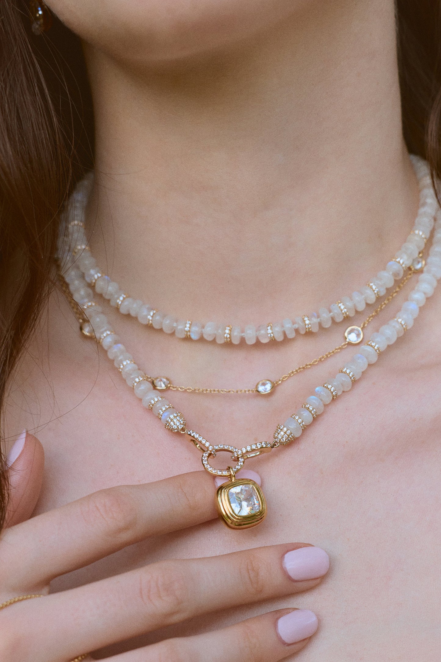 CLOSE UP OF A MODEL’S NECK WEARING A NECKLACE STACK WITH THE BOUJEE BEADS MOONSTONE LONG NECKLACE, AS HER HAND IS LIFTED UP TOUCHING THE NECKLACE PENDANT. 