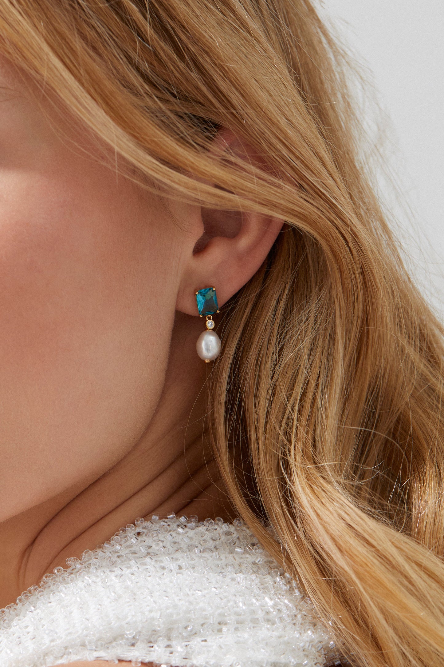 BLUE LAGOON PEARL DROP EARRINGS ON MODEL. CLOSE UP IMAGE OF HER EARS WITH HER FACE CROPPED OUT. SHES WEARINGA. WHITE SEQUIN TOP WITH HER BLONDE HAIR TUCKED BEHIND THE EARS.