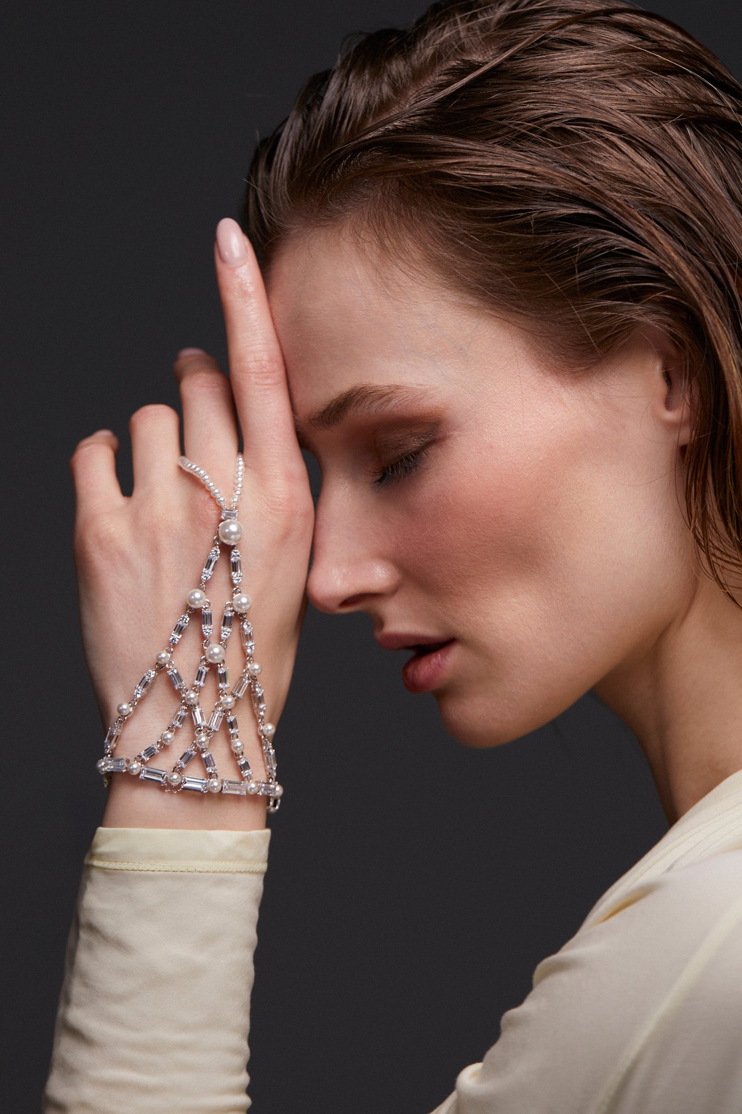 MODEL WEARING INGRID FLOWER HAND CHAIN BRACELET. IT IS A SIDE PROFILE SHOT OF THE MODEL WITH HER HAND UP TO HER FACE. SHE IS WEARING A CREAM SHEER LONG SLEEVE TOP. 