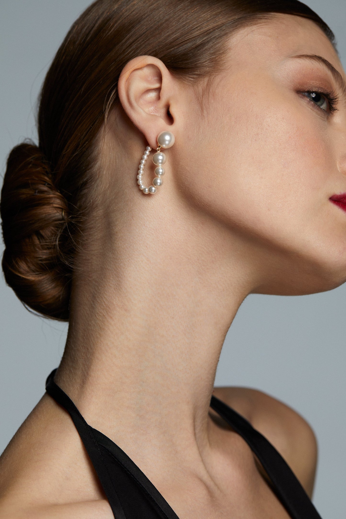 LONDON PEARL DROP C HOOP EARRINGS ON MODEL. HER HAIR IS IN A SLICK BACK BUN. SIDE PROFILE SHOT FOR A SIDE ANGLE OF THE EARRINGS.