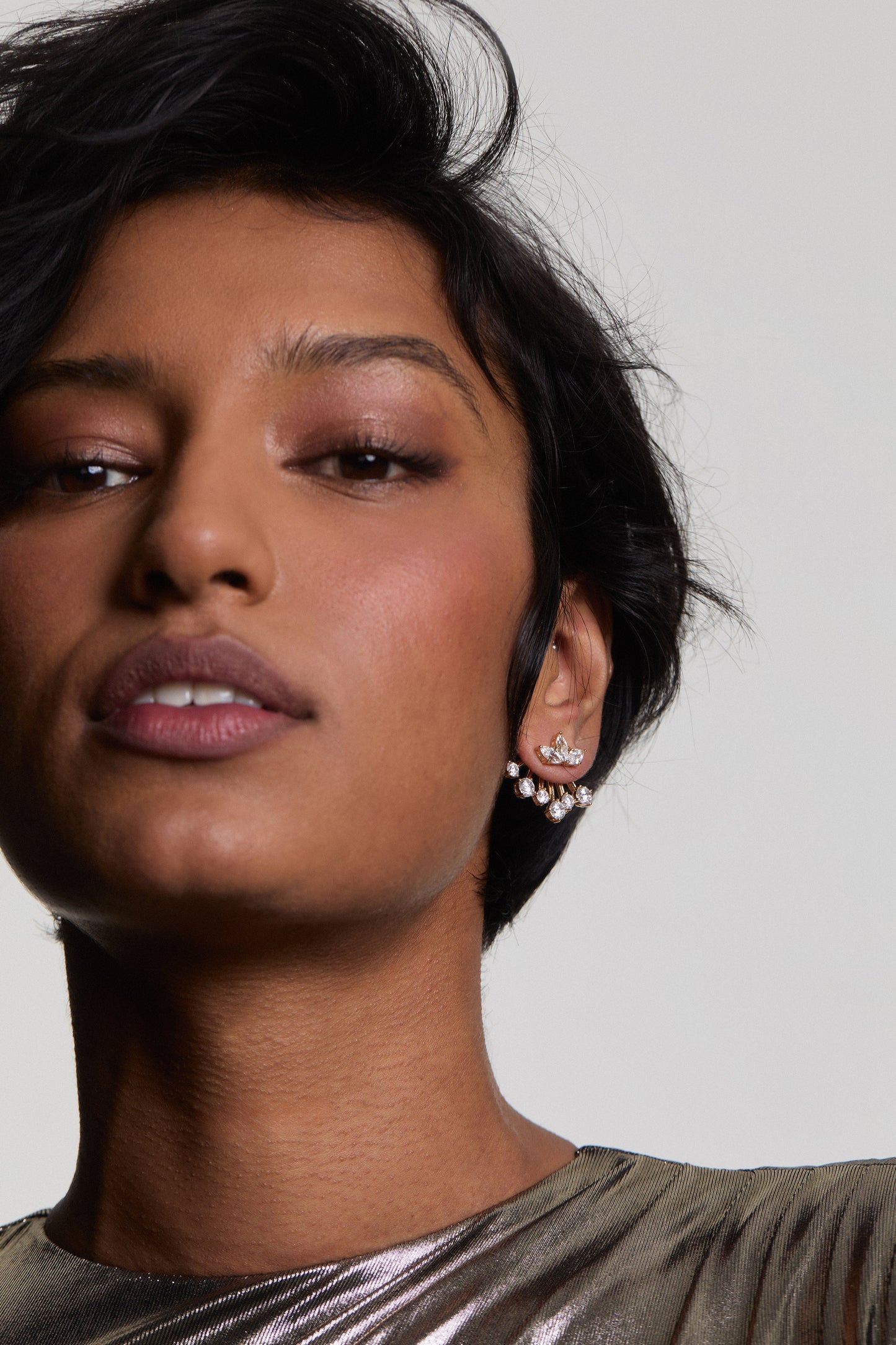 A CLOSE-UP OF A MODEL LOOKING AT THE CAMERA WEARING LUCIA FRONTBACK JACKET EARRINGS, STYLED WITH SHORT HAIR AND A METALLIC TOP