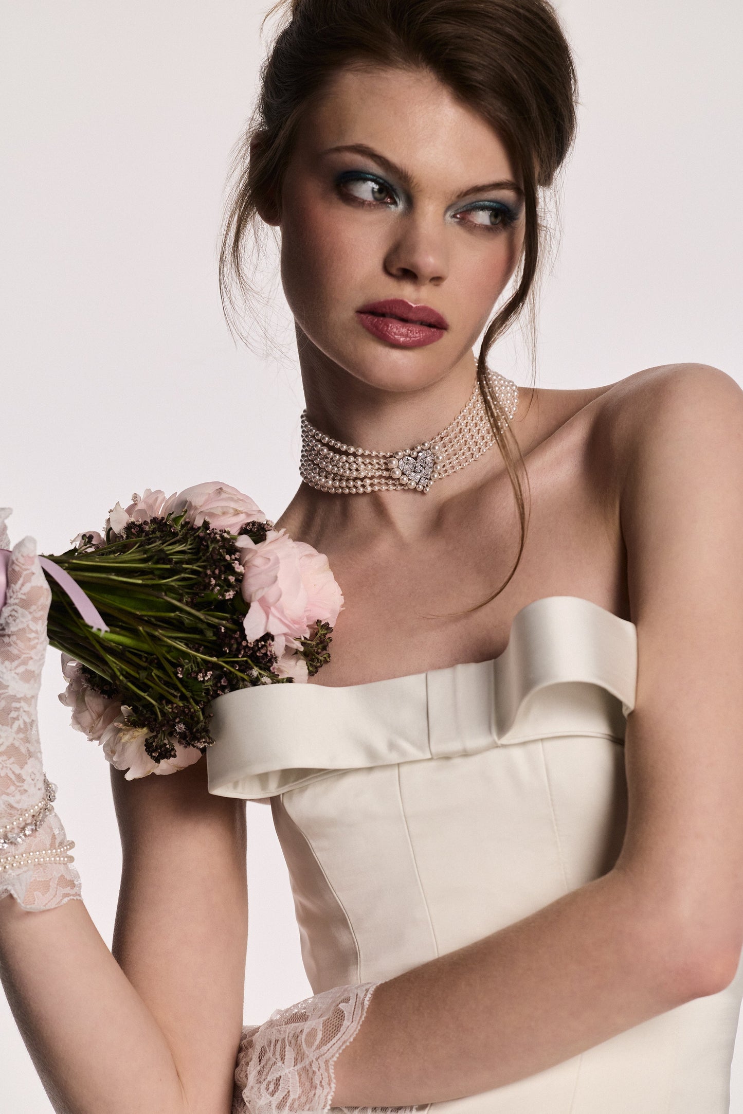 A MODEL LOOKING AWAY FROM THE CAMERA, HOLDING UP A BOUQUET OF FLOWERS TO HER SHOULDER WHILE WEARING THE MATERIAL PEARL CHOKER NECKLACE AND WHITE LACE GLOVES.