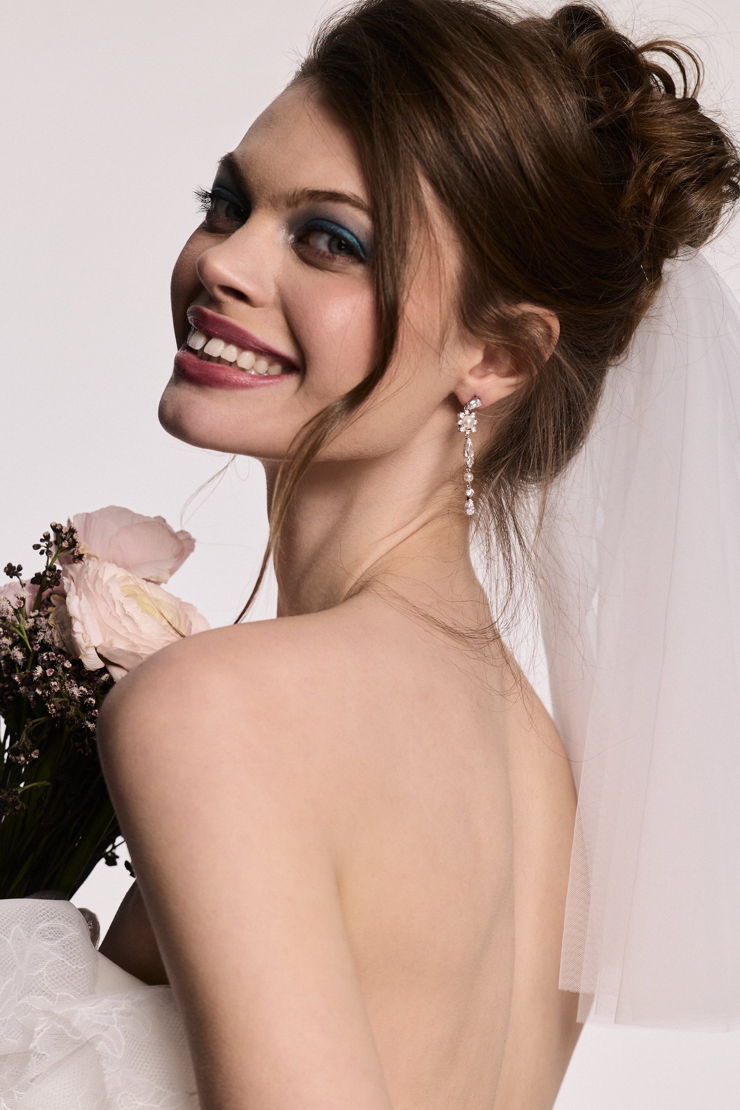 A MODEL LOOKING OVER HER SHOULDER WITH HER BACK TO THE CAMERA, WEARING THE MATERIAL PEARL LINEAR EARRINGS, A VEIL IN HER HAIR, AND HOLDING A BOUQUET OF FLOWERS. 