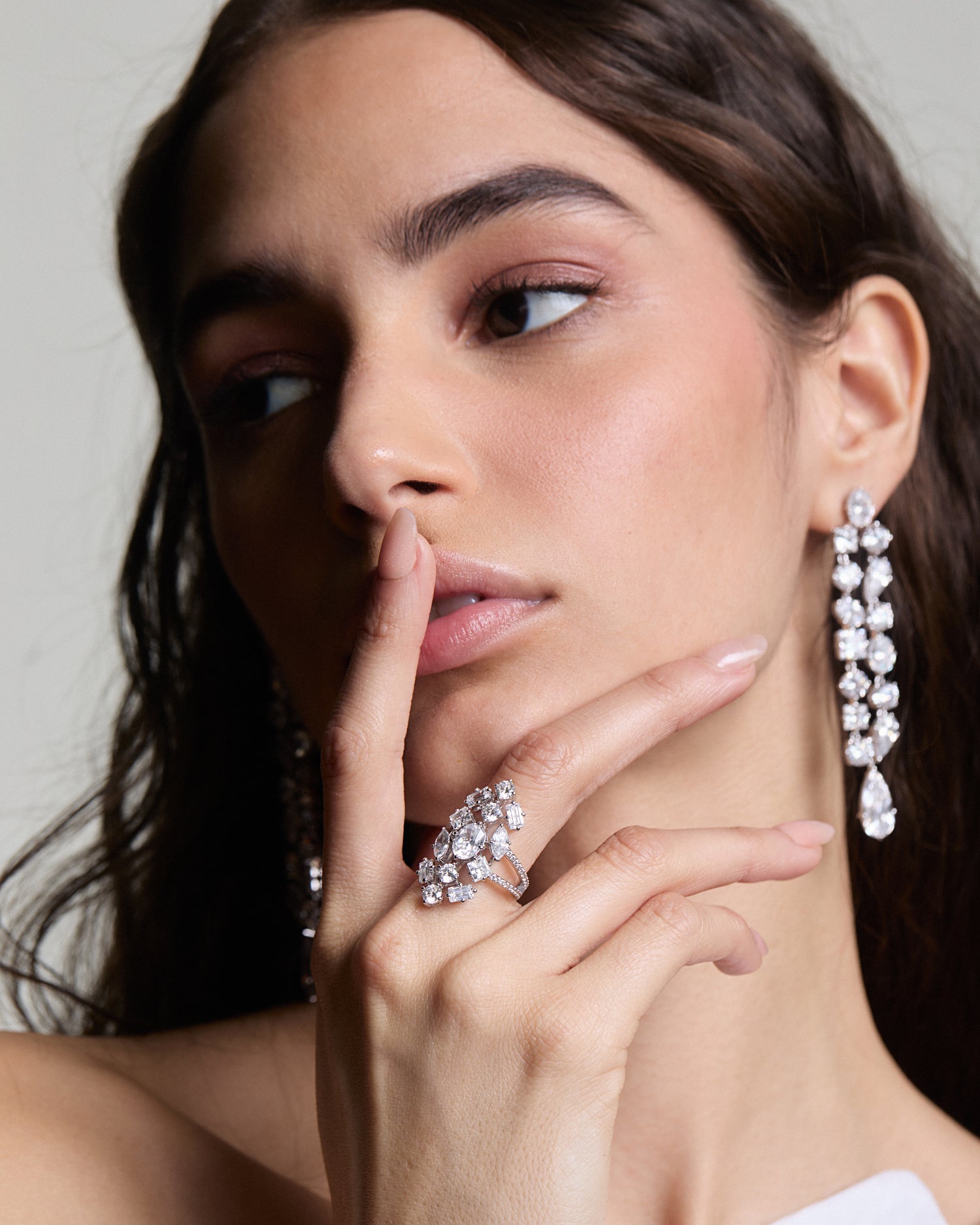 Image of model wearing Nadri's Riviera CZ ring and Double Linear earrings.  She is looking to the side with one finger on her lips and her dark hair is falling in loose waves.