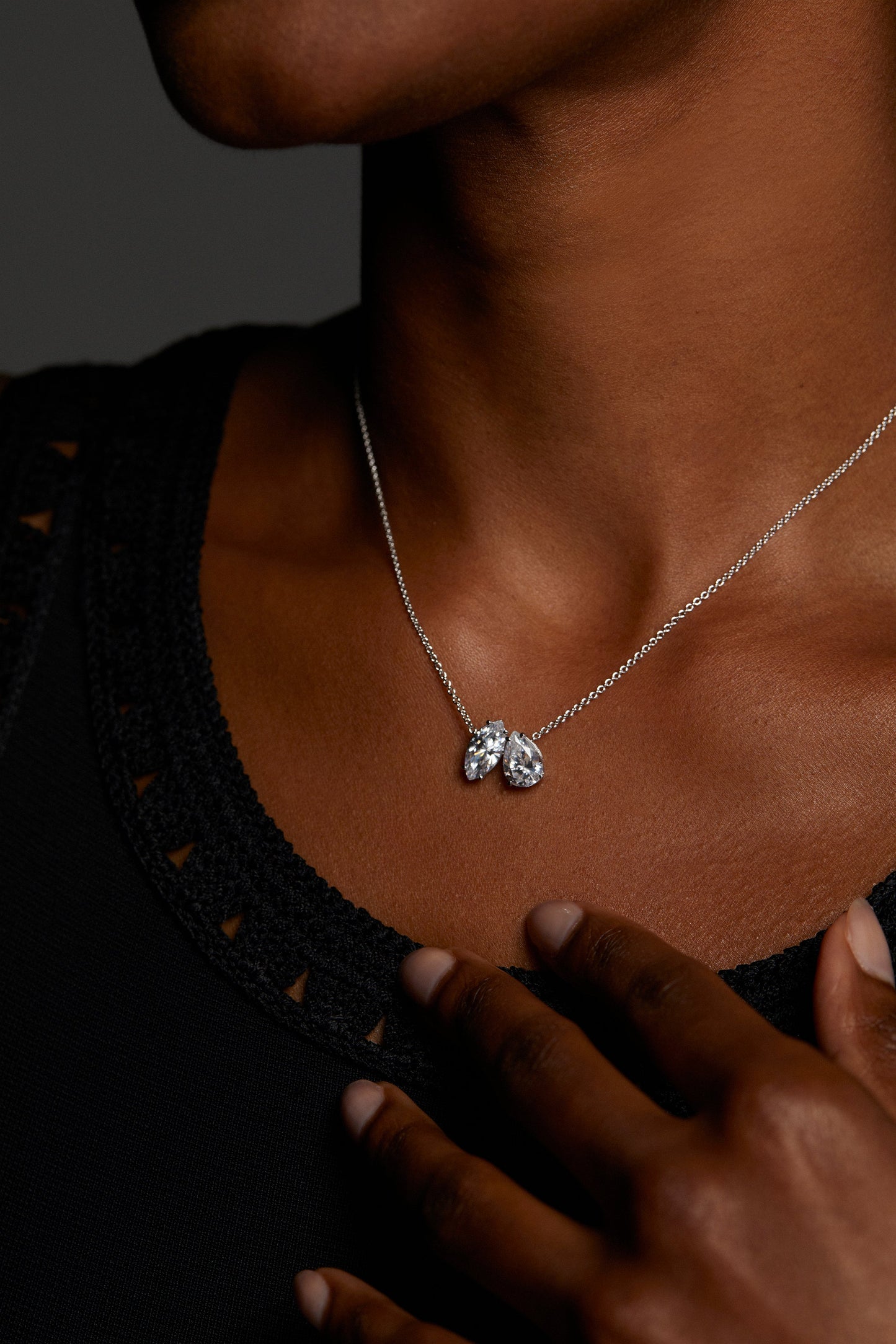 MODEL WEARING EVERLEIGH PENDANT NECKLACE. IT IS A CLOSE UP IMAGE OF HER NECKLINE. HER HAND IS REACHING TOWARDS THE NECKLACE. SHE IS WEARING A BLACK CROCHET TOP. 
