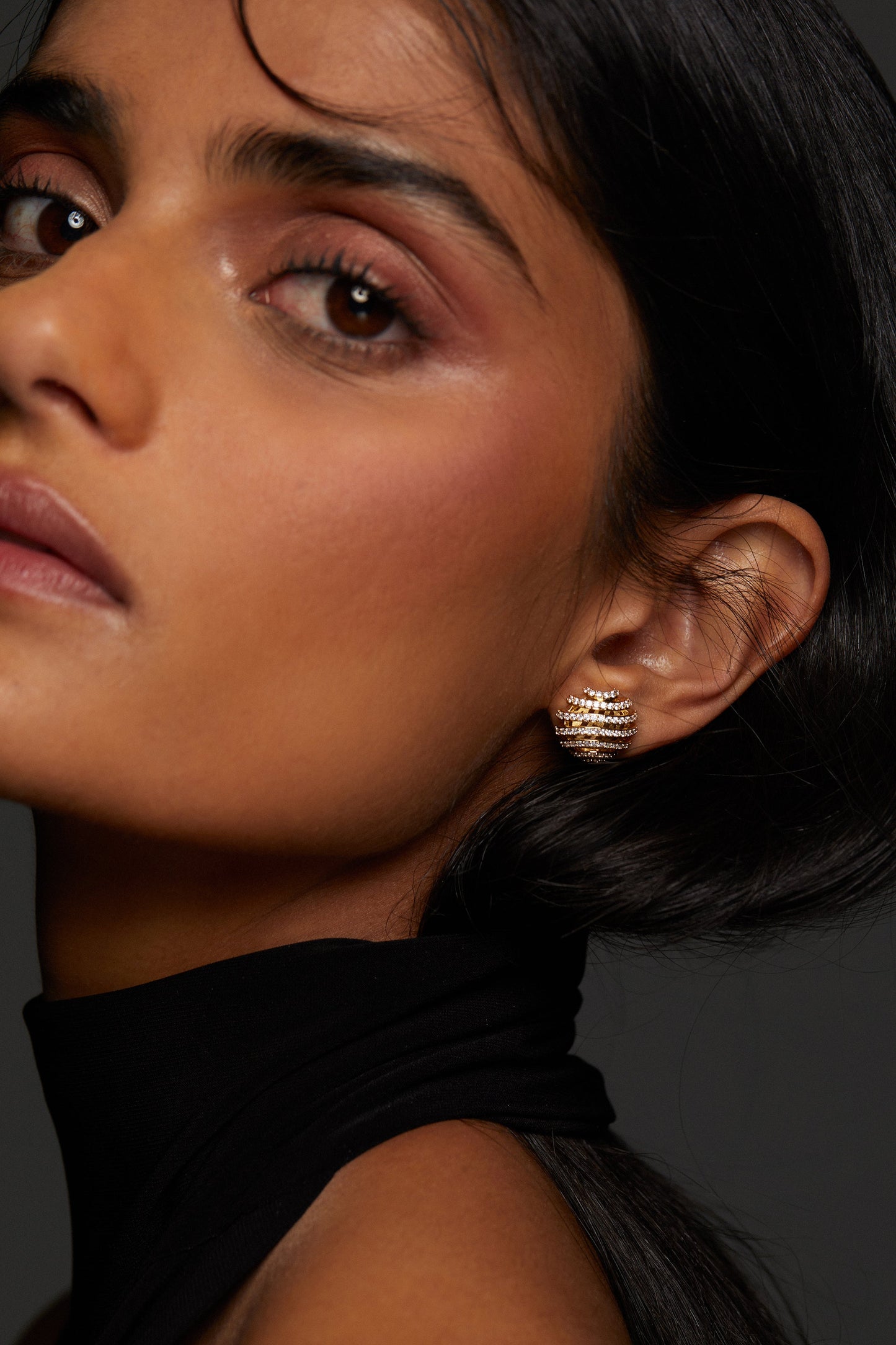 CLOSE UP IMAGE OF MODEL WEARING PISA OPEN PAVE STUD EARRINGS. SHE IS LOOKING DIRECTLY INTO THE CAMERA. HER HAIR IS TUCKED BEHIND HER EAR AND SHE IS WEARING A BLACK HIGH NECK SLEEVELESS TOP. 