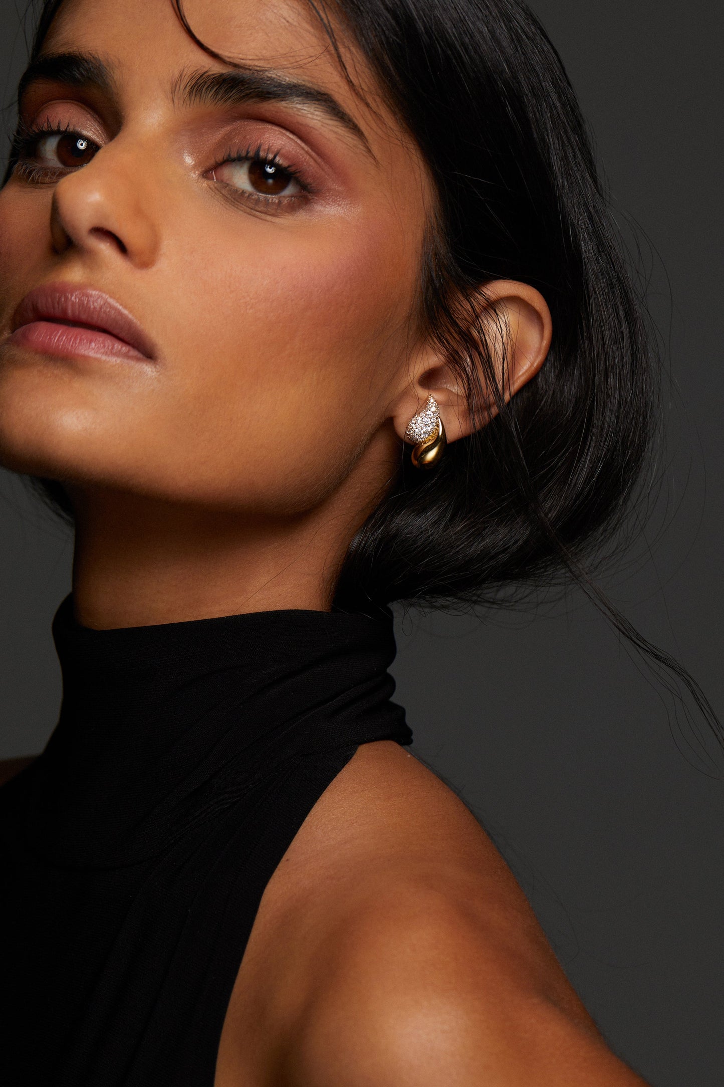 CLOSE UP OF MODEL WEARING PISA DOUBLE TEARDROP EARRINGS WITH HER HAIR TUCKED BEHIND HER EAR. SHE IS LOOKING DIRECTLY INTO THE CAMERA WITH A BLACK HIGH NECK SLEEVELESS TOP. 