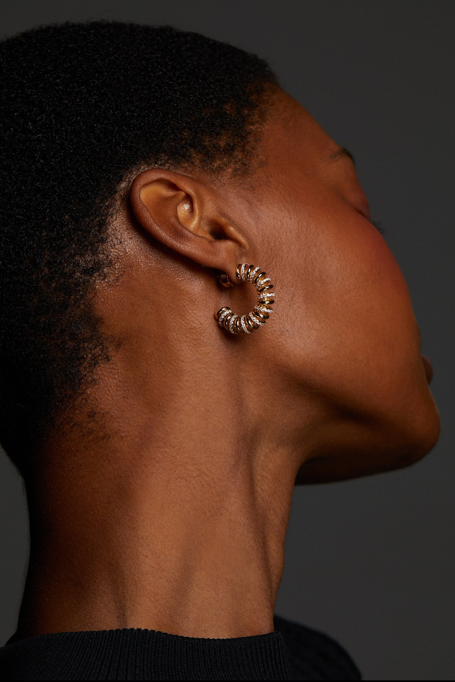 MODEL WEARING PISA COIL C HOOP EARRINGS. SHE IS FACING AWAY FROM THE CAMERA AND WEARING A BLACK HIGH NECK TOP. 
