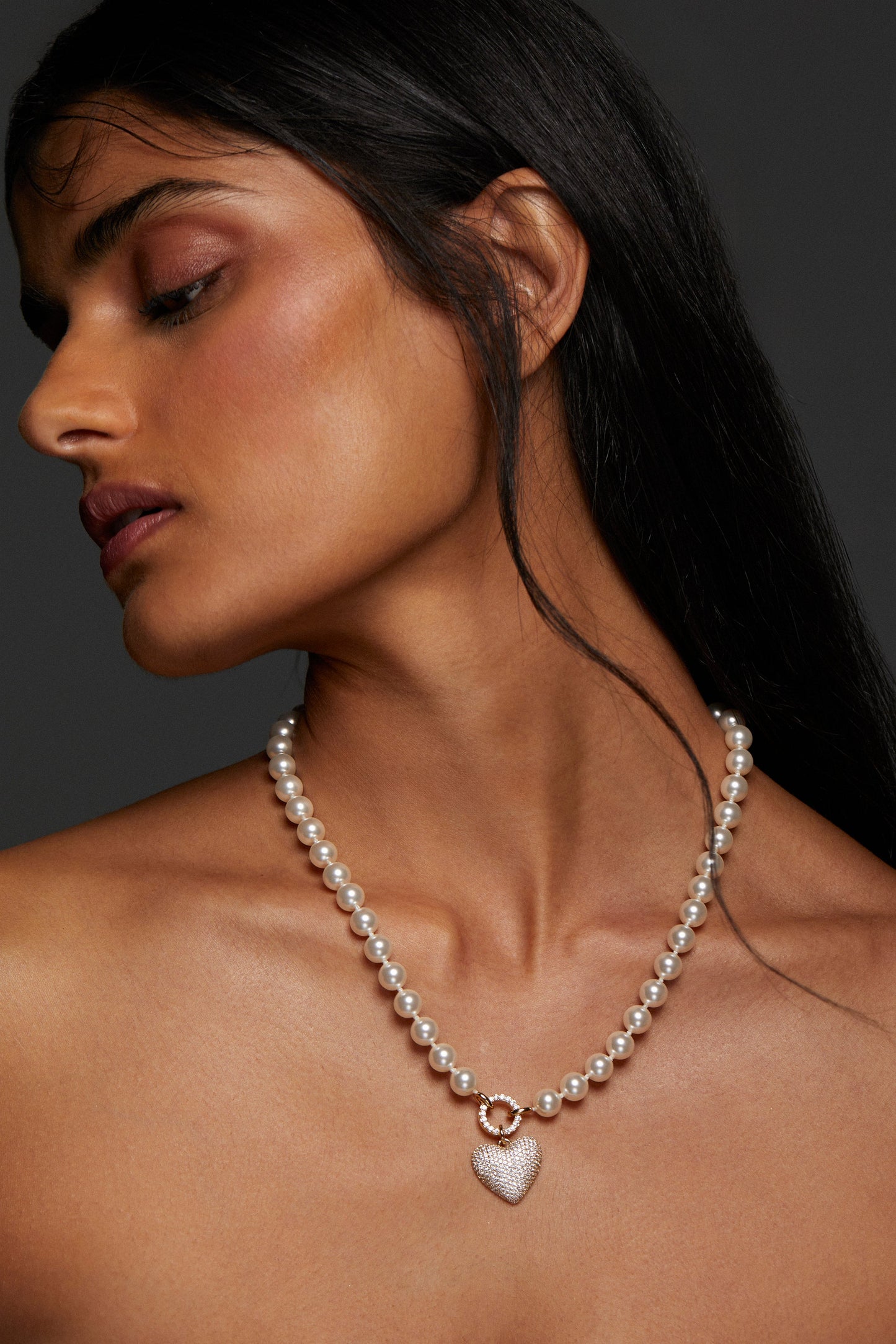 MODEL WEARING POPPY 8MM CZ HEART NECKLACE. IT IS A CLOSE UP IMAGE OF HER NECKLINE - SHE IS LOOKING DOWN WITH HER EYES ALMOST CLOSED. HER LONG HER IS BEHIND HER SHOULDERS. 