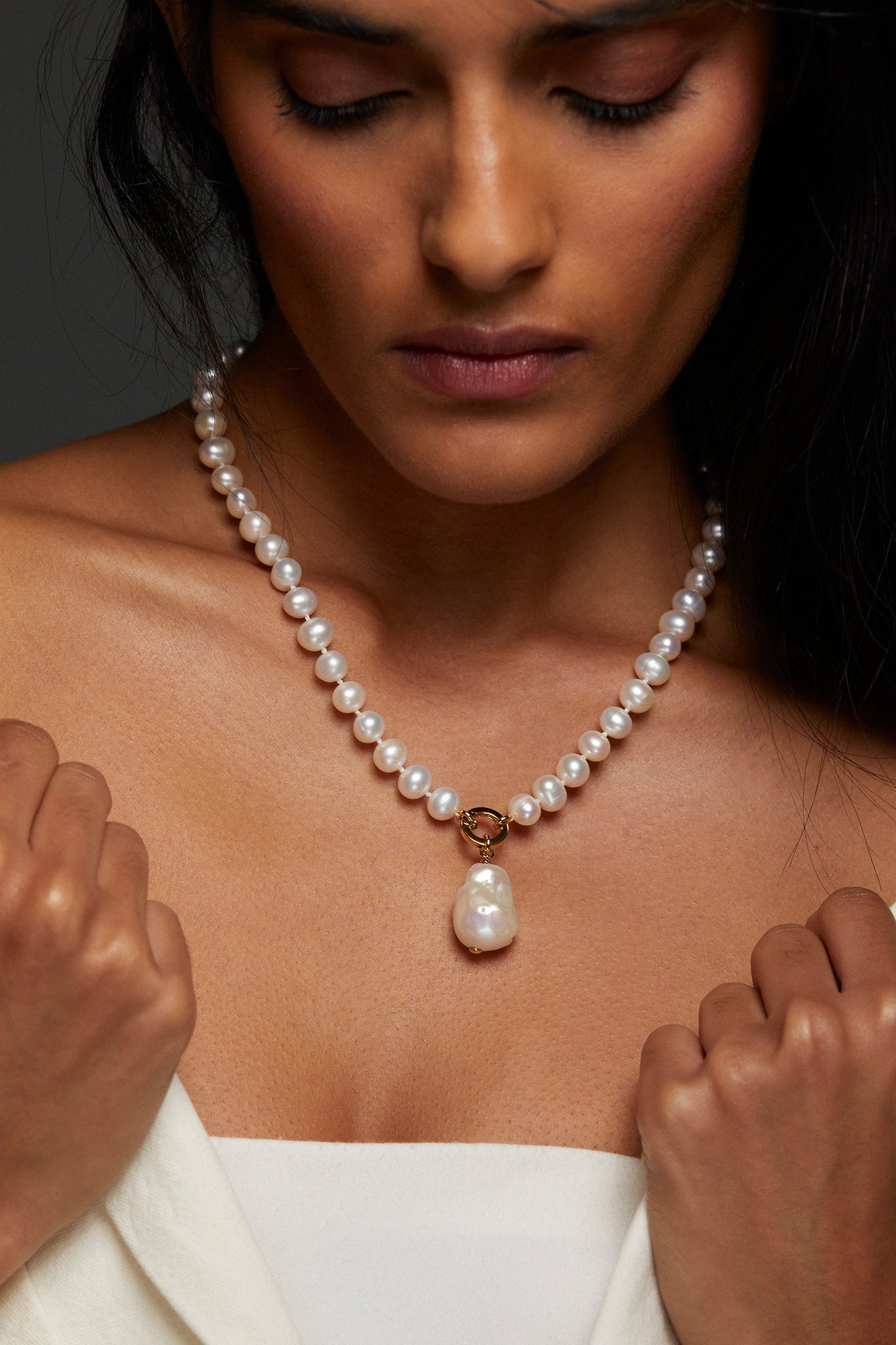MODEL WEARING POPPY 8MM BOLD PEARL CHARM NECKLACE. SHE IS LOOKING DOWN WITH HER EYES CLOSED AND HAS ON A STRAPLESS WHITE TOP WITH A BLAZER OVER IT THAT SHE IS HOLDING WITH BOTH HANDS. HER HAIR IS PULLED BACK BEHIND HER SHOULDERS. 
