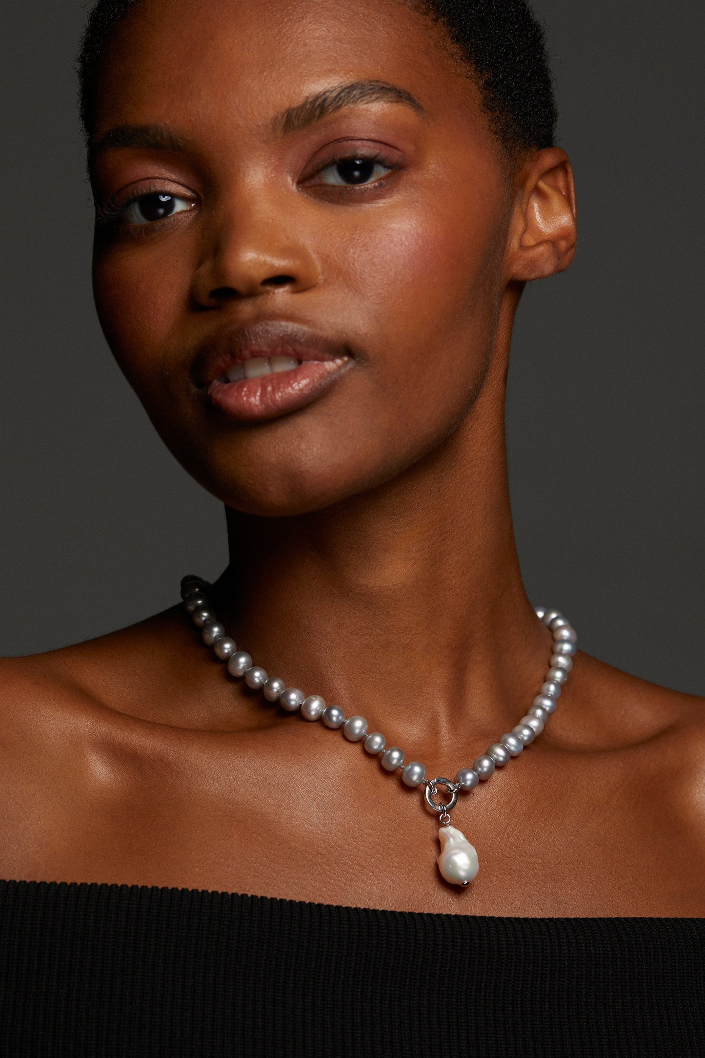 MODEL WEARING POPPY 8MM GRAY PEARL CHARM NECKLACE. SHE IS LOOKING DIRECTLY INTO THE CAMERA AND IT IS A CLOSE UP SHOWING HER NECKLINE. SHE IS WEARING AN OFF THE SHOULDER BLACK TOP. 