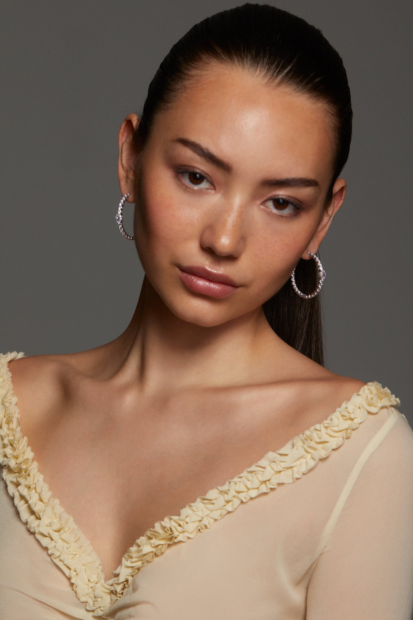 Model wearing a sheer beige top with floral details against a gray background. She is looking directly into the camera with her hair pulled back into a ponytail wearing STELLAR CZ HOOP EARRINGS