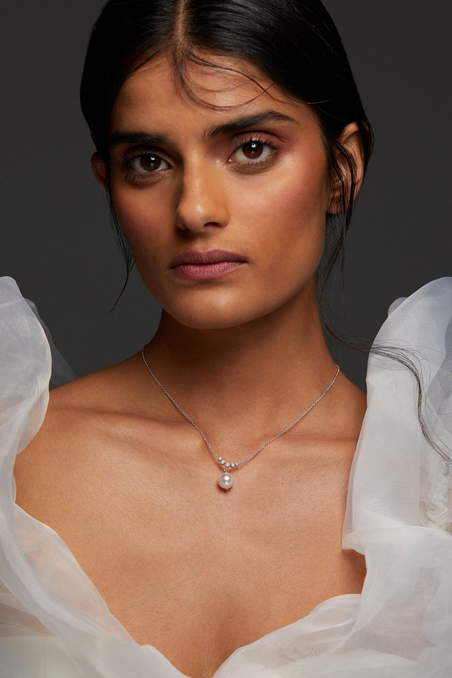 MODEL WEARING VENUS PEARL DROP NECKLACE. SHE IS LOOKING DIRECTLY INTO THE CAMERA WITH HER HAIR BACK IN A LOW BUN WEARING PUFFY WHITE SLEEVE DRESS. 