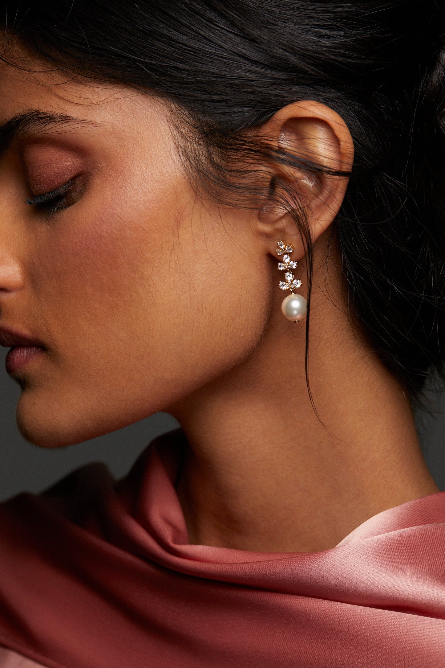 MODEL WEARING VENUS SHORT PEARL DROP EARRINGS CLOSE UP WITH HER HAIR IN A LOW BUN AND A PINK HIGH NECK CHIFFON TOP. 