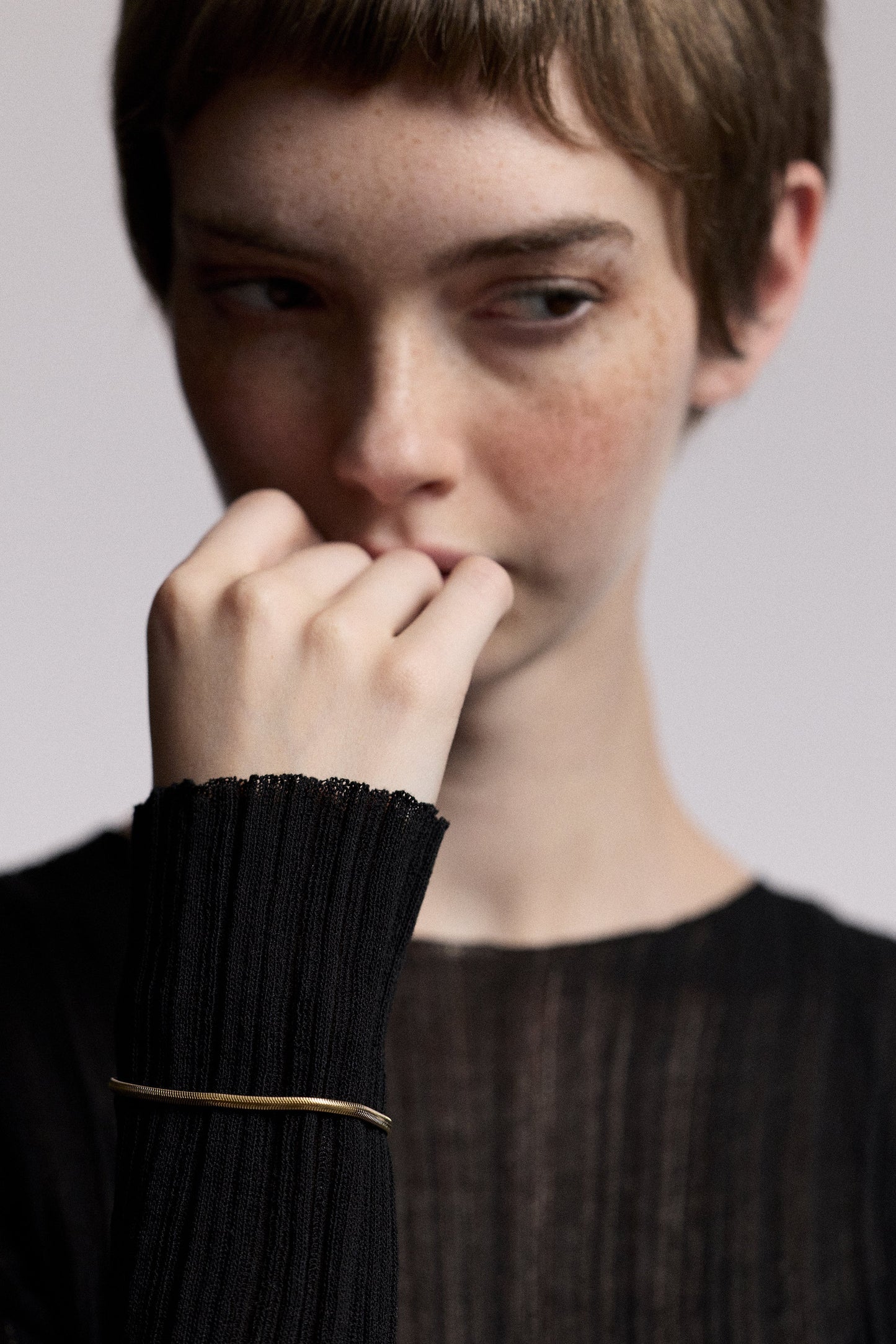 A MODEL LOOKING OFF TO THE SIDE WITH HER HAND LIFTED IN FRONT OF HER AGAINST HER MOUTH, WEARING A SHEER BLACK DRESS AND THE CELINE SNAKE CHAIN BRACELET