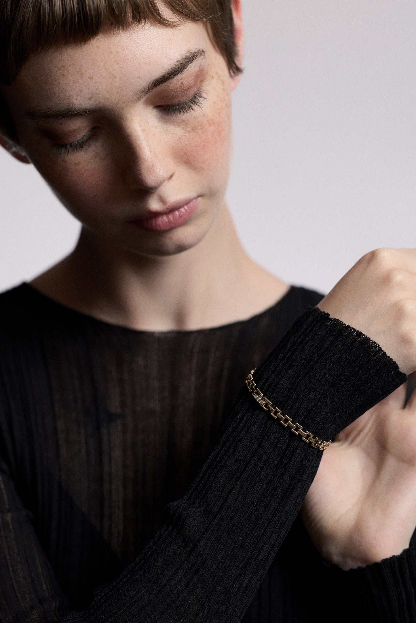 A MODEL LOOKING DOWN AT HER HAND RAISED IN FRONT OF HER, WEARING A SHEER BLACK DRESS AND THE CELINE SMALL WATCH CHAIN BRACELET