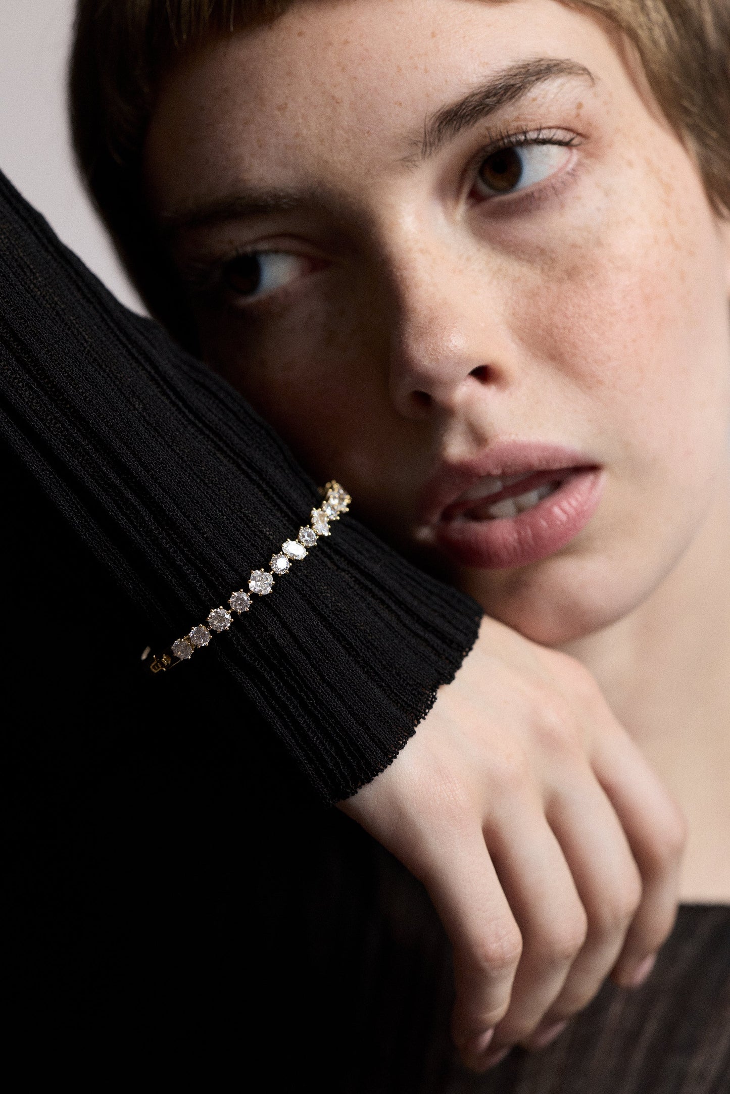 CLOSE UP OF A MODEL’S FACE AS SHE LIFTS UP HER HAND NEXT TO HER CHEEK, WEARING A SHEER BLACK DRESS AND THE CELINE MULTI CZ HINGE BRACELET