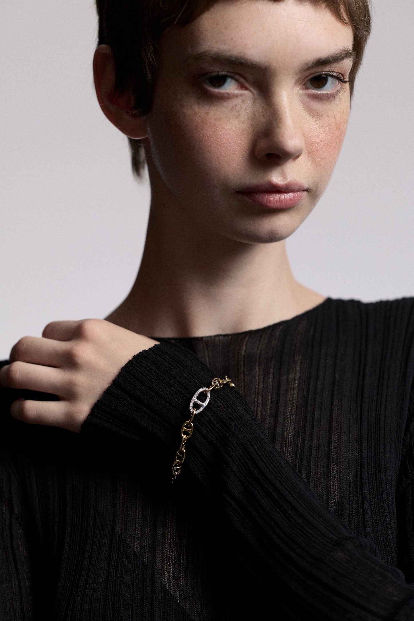 A MODEL LOOKING AT THE CAMERA WITH HER HAND RESTING ON THE OPPOSITE SHOULDER, AS SHE WEARS A SHEER BLACK DRESS AND THE CELINE ANCHOR LINK BRACELET