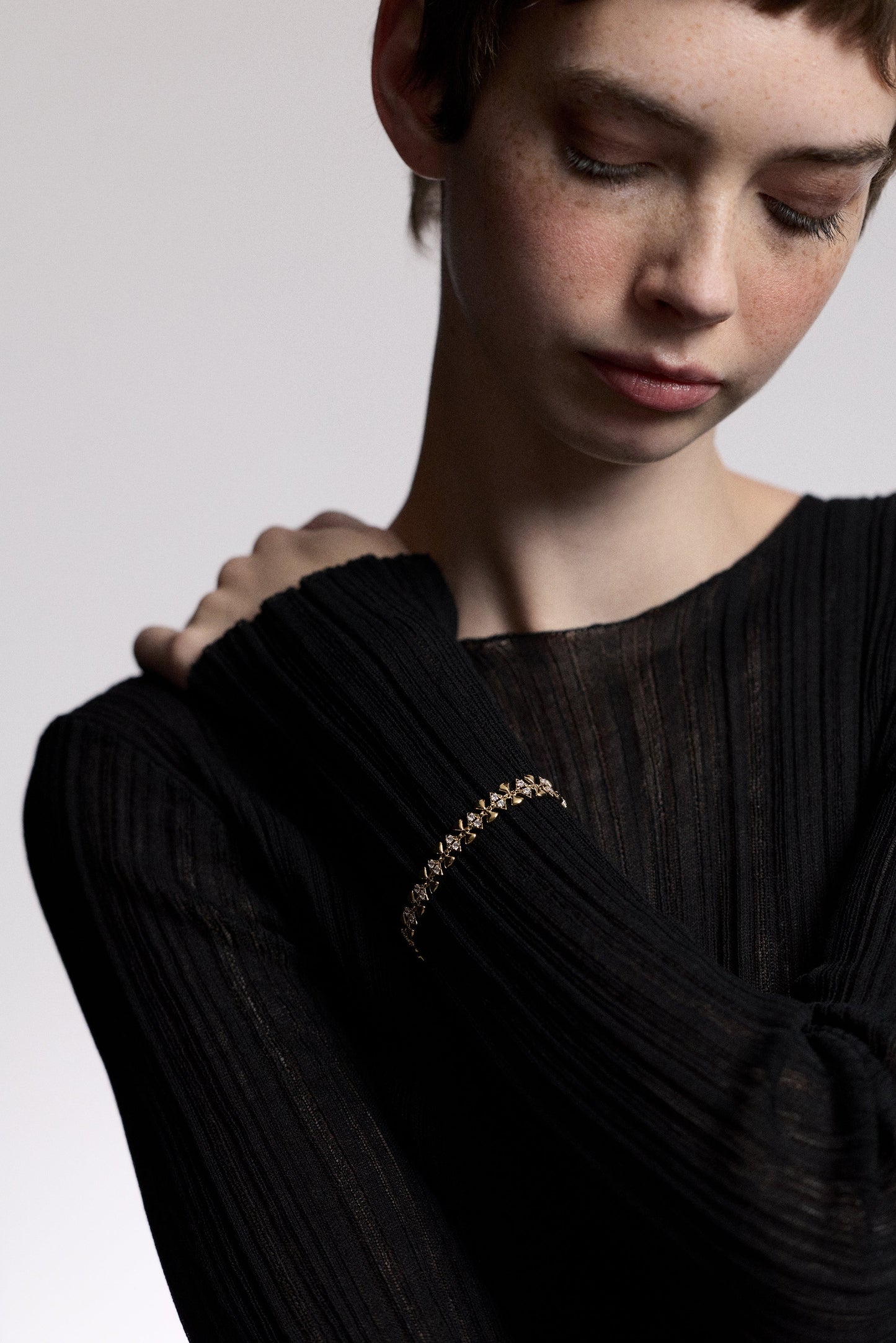 A MODEL LOOKING DOWN WITH HER HAND RESTING AGAINST HER OPPOSITE SHOULDER, AS SHE WEARS A SHEER BLACK DRESS AND THE CELINE GEO LINE BRACELET