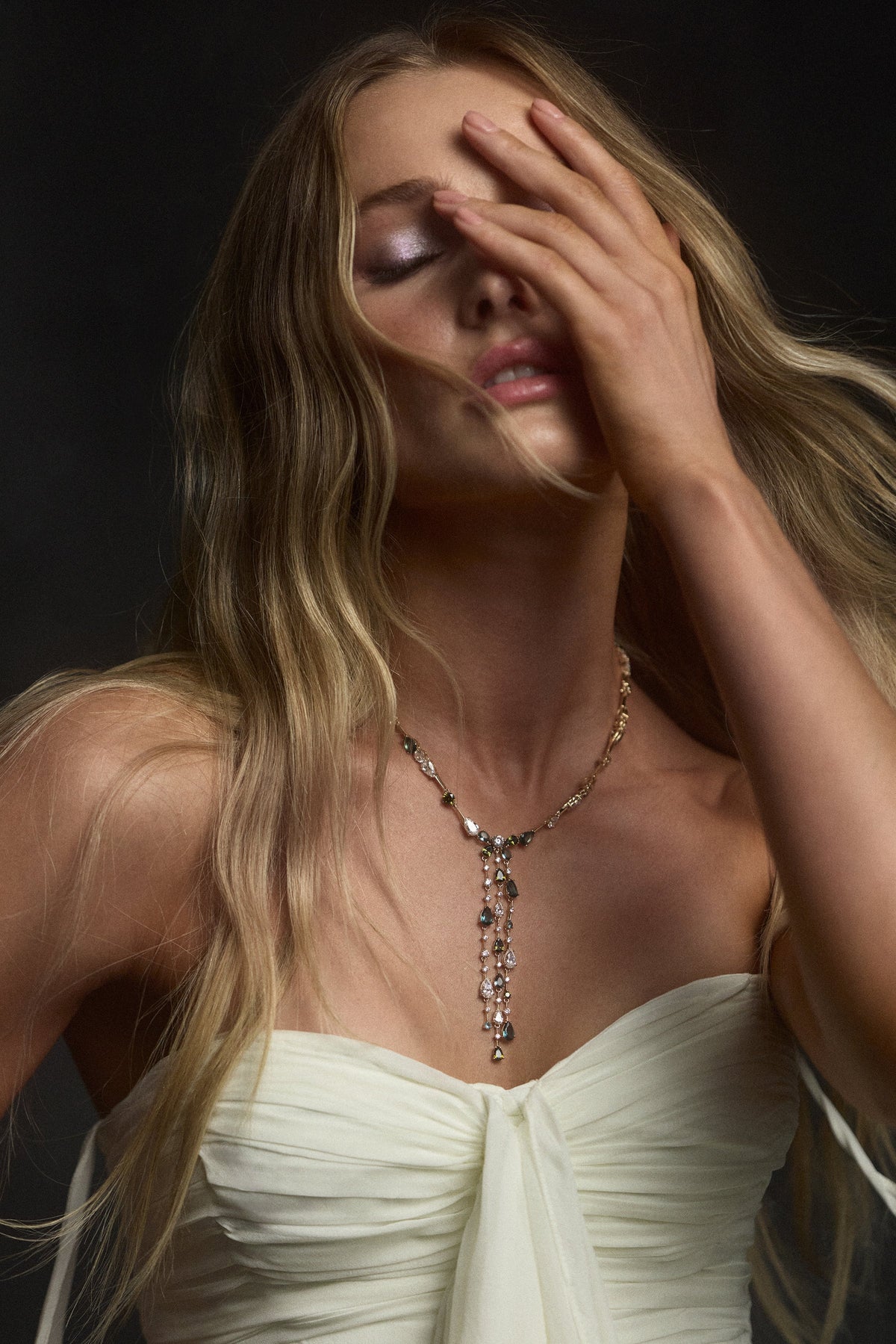 A MODEL WITH HER EYES CLOSED AND HER HAND LIFTED UP TO HER FACE, WEARING A STRAPLESS WHITE DRESS AND THE HERA MULTI DRAMA Y NECKLACE