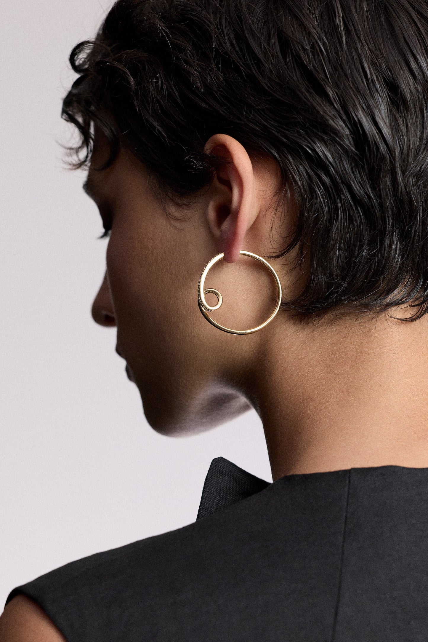 THE BLACK OF A MODEL’S HEAD TURNED SLIGHTLY TO THE SIDE, WITH SHORT BLACK HAIR WEARING THE MUSE SWIRL HOOP EARRINGS