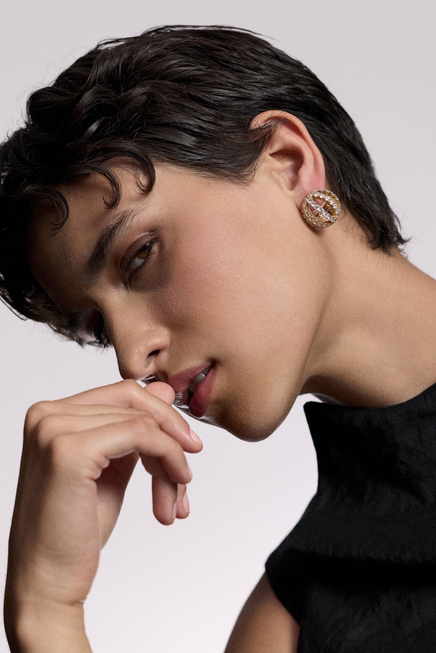 A MODEL TURNED SLIGHTLY TO THE SIDE LOOKING AT THE CAMERA WITH HER HAND LIFTED UP TO HER MOUTH, AS SHE WEARS A BLACK TOP AND THE MUSE SPHERE BUTTON EARRINGS