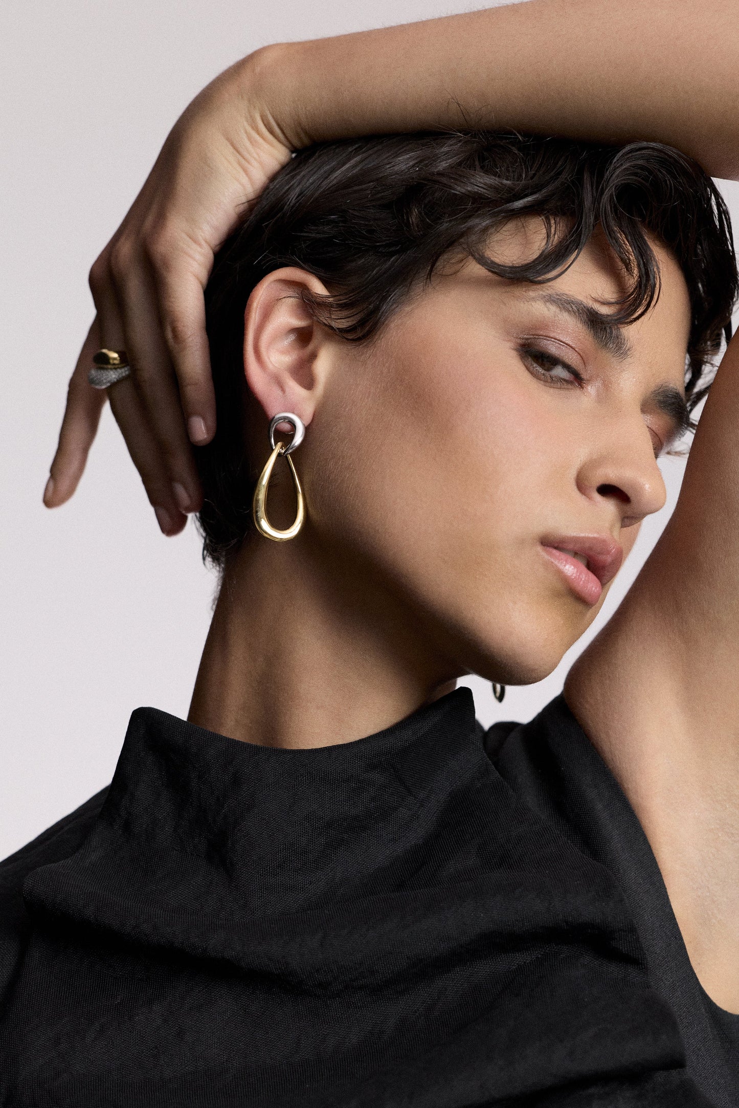 A MODEL TURNED SLIGHTLY TO THE SIDE WITH HER ARM RAISED AND DRAPED OVER HER HEAD, AS SHE WEARS A BLACK TOP AND THE MUSE DOOR KNOCK EAR EARRINGS