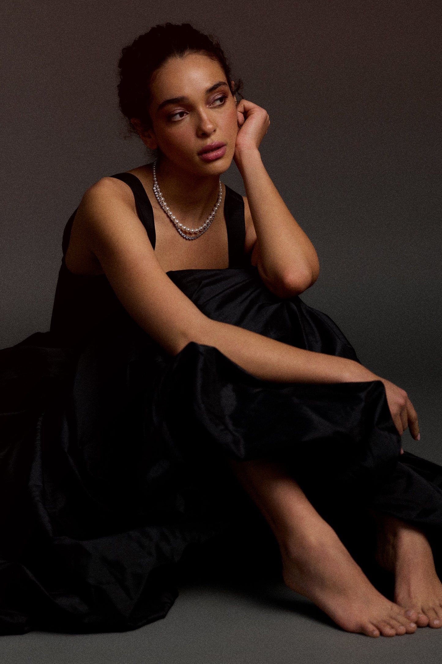 MODEL SITTING ON THE FLOOR IN A BLACK DRESS WITH HER LEGS PULLED TO HER CHEST, LOOKING AWAY WITH ONE ARM RESTING ON HER KNEES AND THE OTHER AGAINST HER HEAD, WEARING THE UNDER THE SEA DOUBLE LAYER NECKLACE.