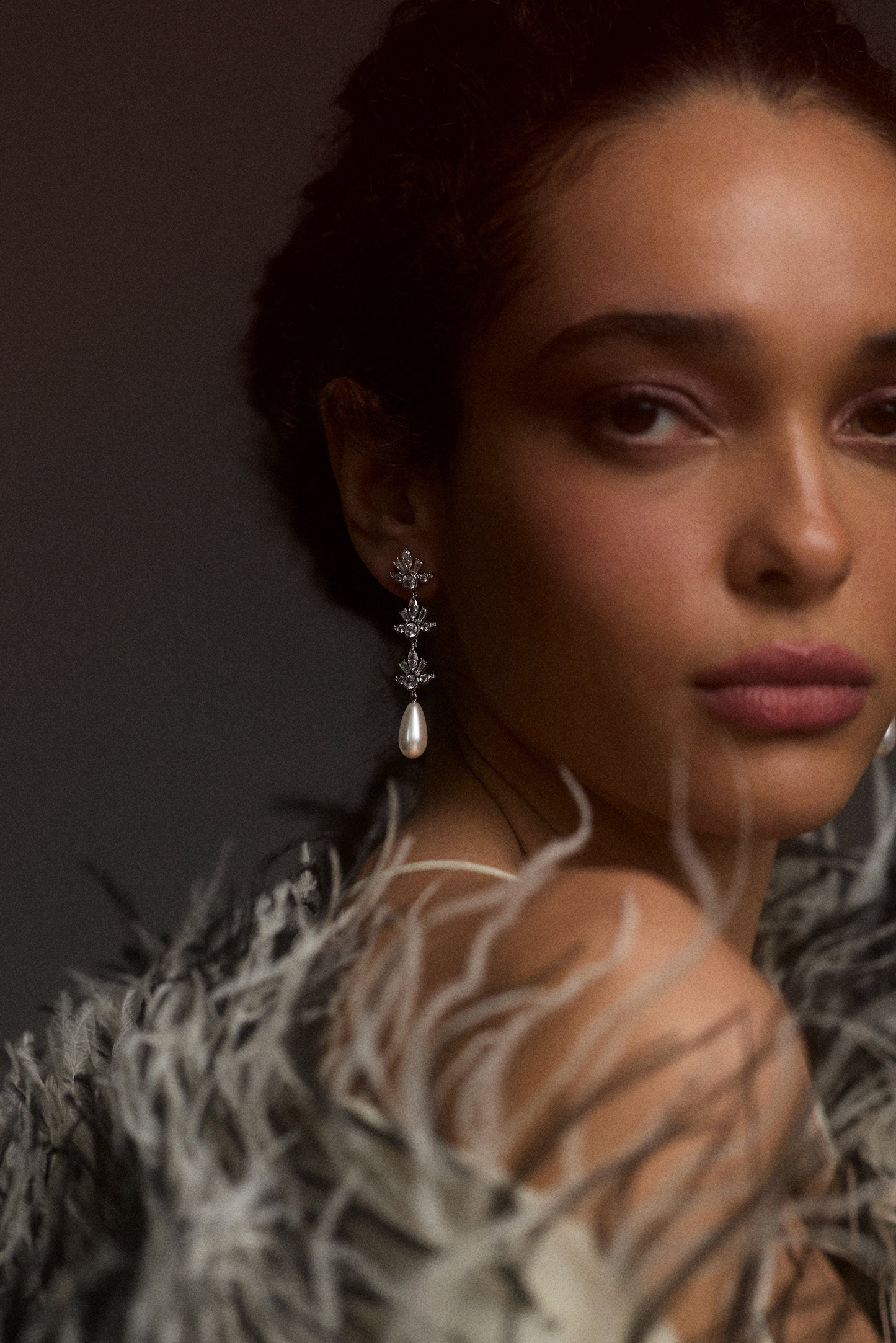 A MODEL LOOKING AT THE CAMERA OVER HER SHOULDER, WITH HER HAIR IN A BUN AND WEARING THE UNDER THE SEA PEARL TRIPLE DROP EARRINGS STYLED WITH A FUR JACKET