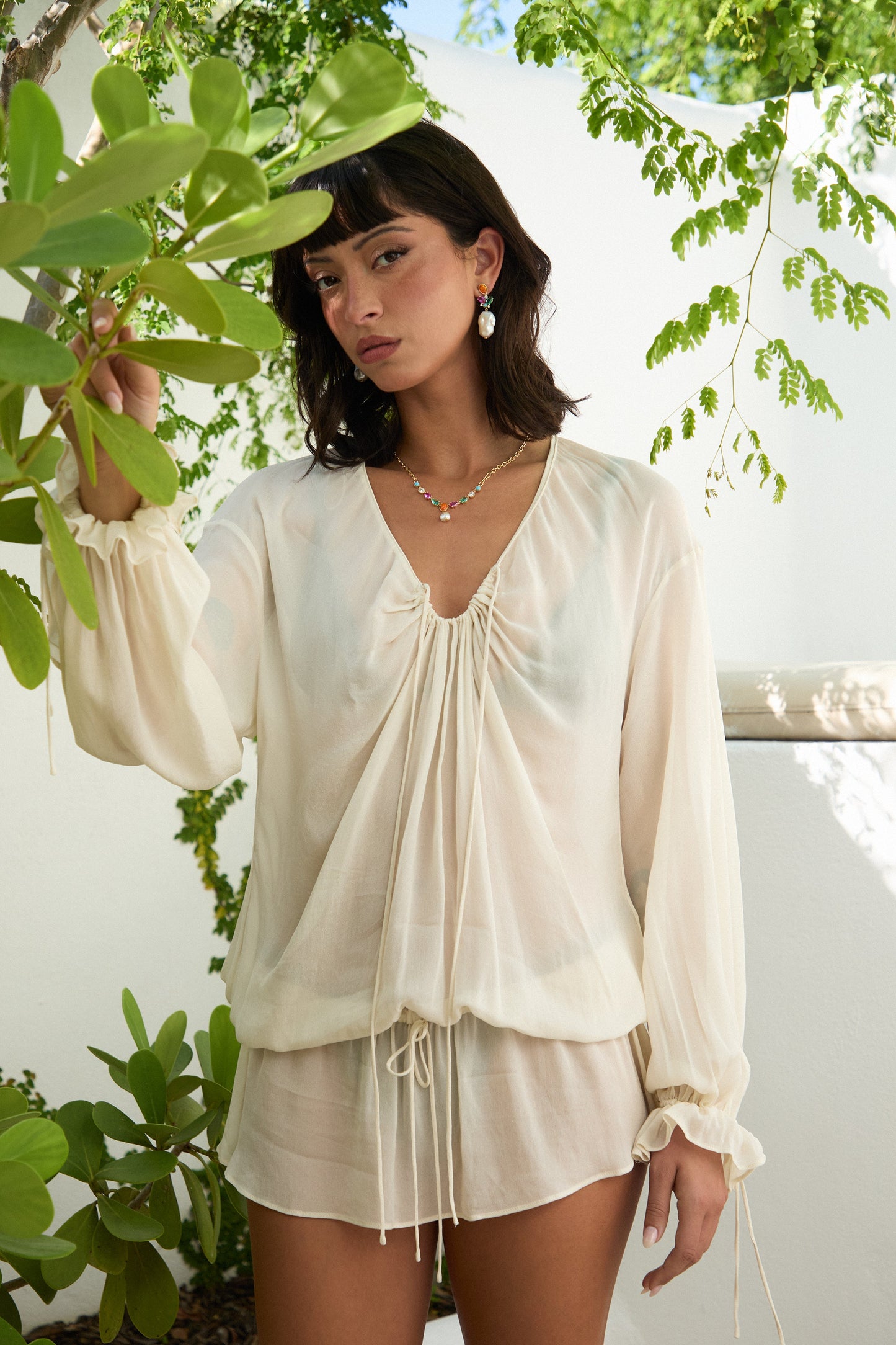 A MODEL LOOKING AT THE CAMERA WITH HER HAND LIFTED GRABBING A LEAFY GREEN PLANT, AS SHE WEARS THE FRIEDA MULTI PEARL FRONTAL NECKLACE AND PEARL DROP EARRINGS