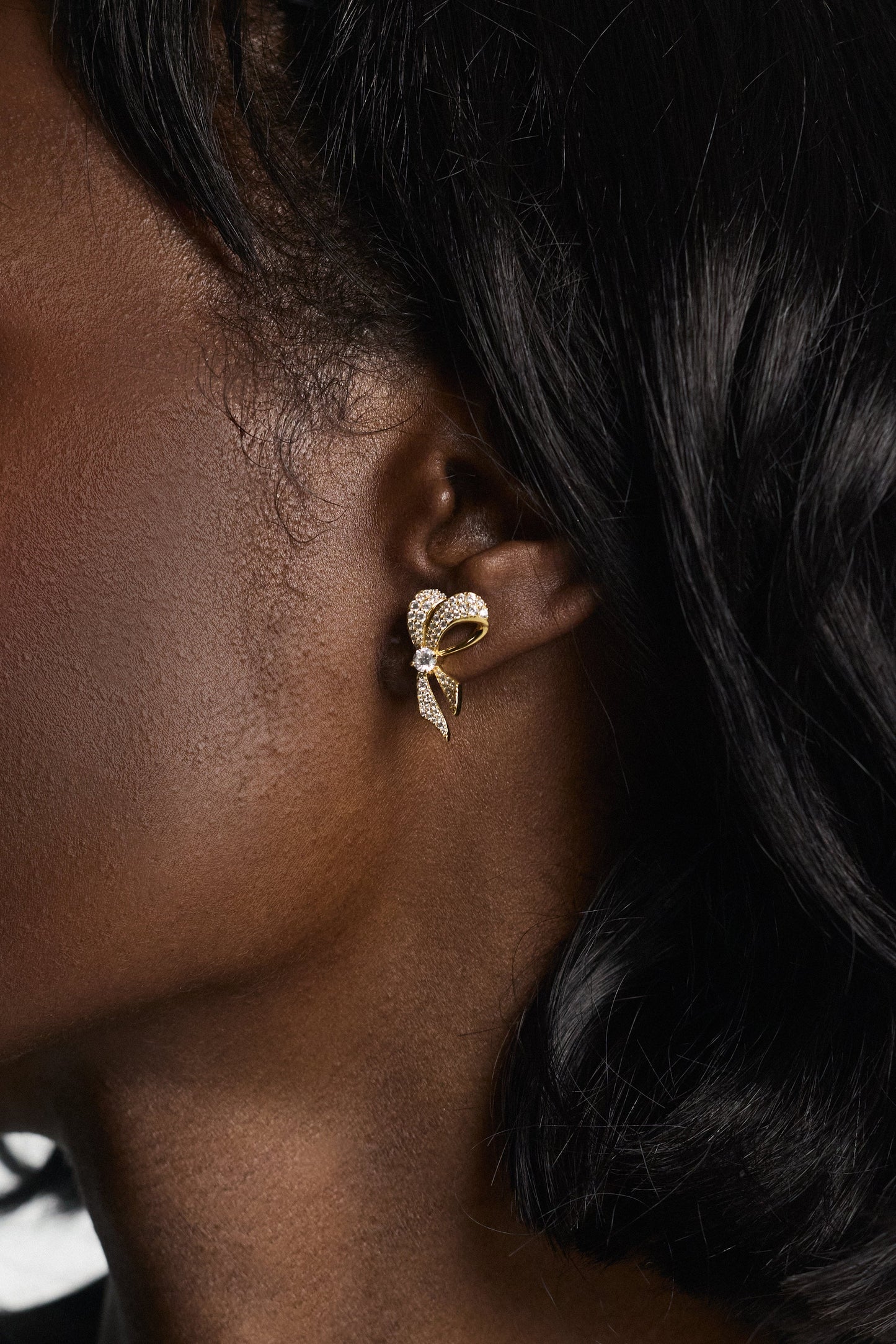 CLOSE UP OF A MODEL’S EAR WEARING THE EROS SMALL RIBBON STUD EARRINGS AND HER BLACK HAIR SWEPT BACK