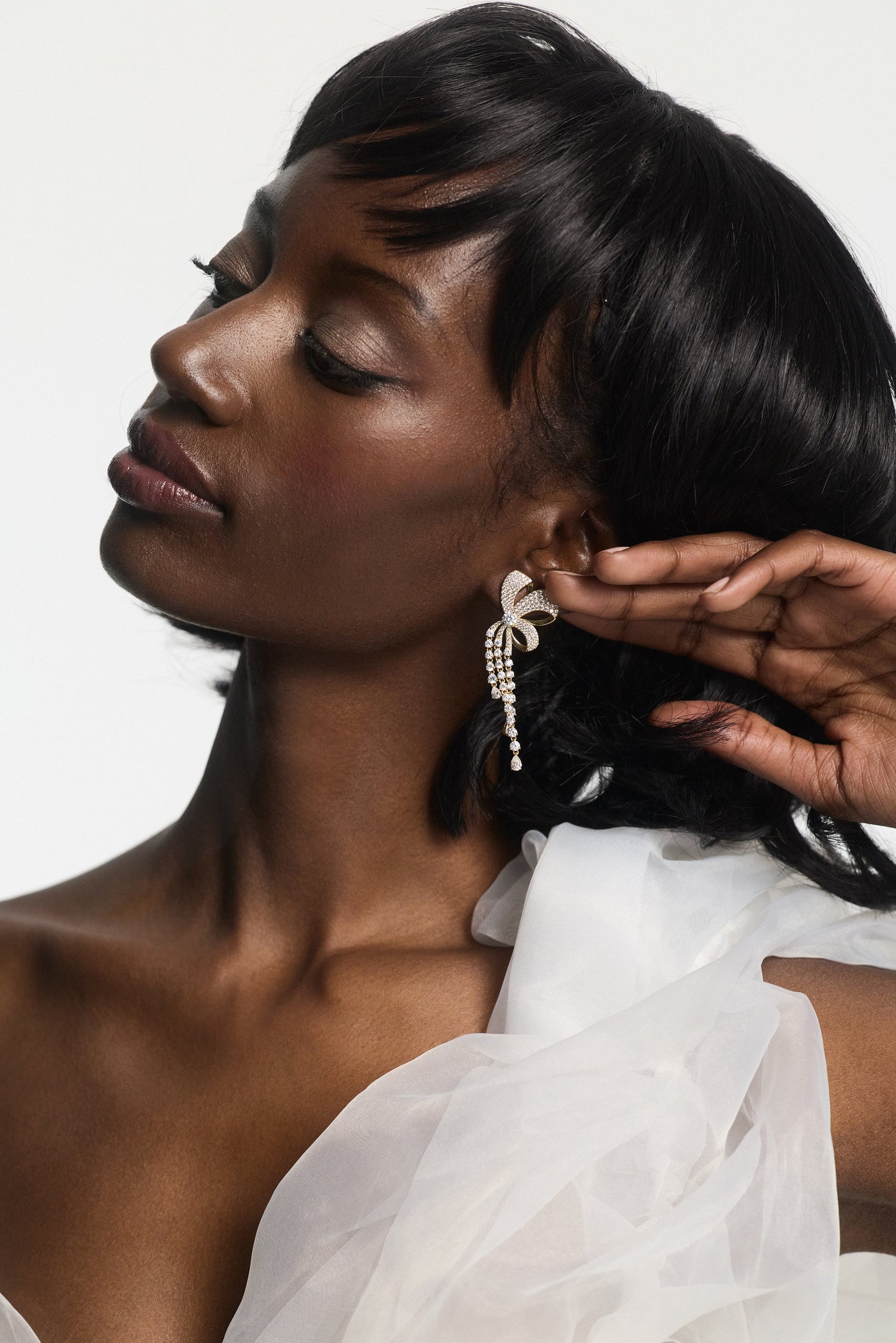 A MODEL LOOKING TO THE SIDE AND SWEEPING HER HAIR BACK, WEARING THE EROS RIBBON CHANDELIER EARRINGS AND A WHITE DRESS WITH CHIFFON SLEEVES