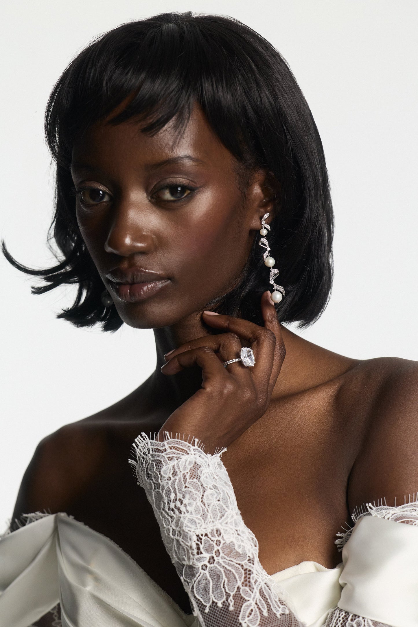 MODEL WEARING A WHITE LACE DRESS WITH EROS RIBBON PEARL LINEAR EARRINGS AND AN ENGAGEMENT RING, ARM FOLDED WITH HAND LIFTED TO HER FACE