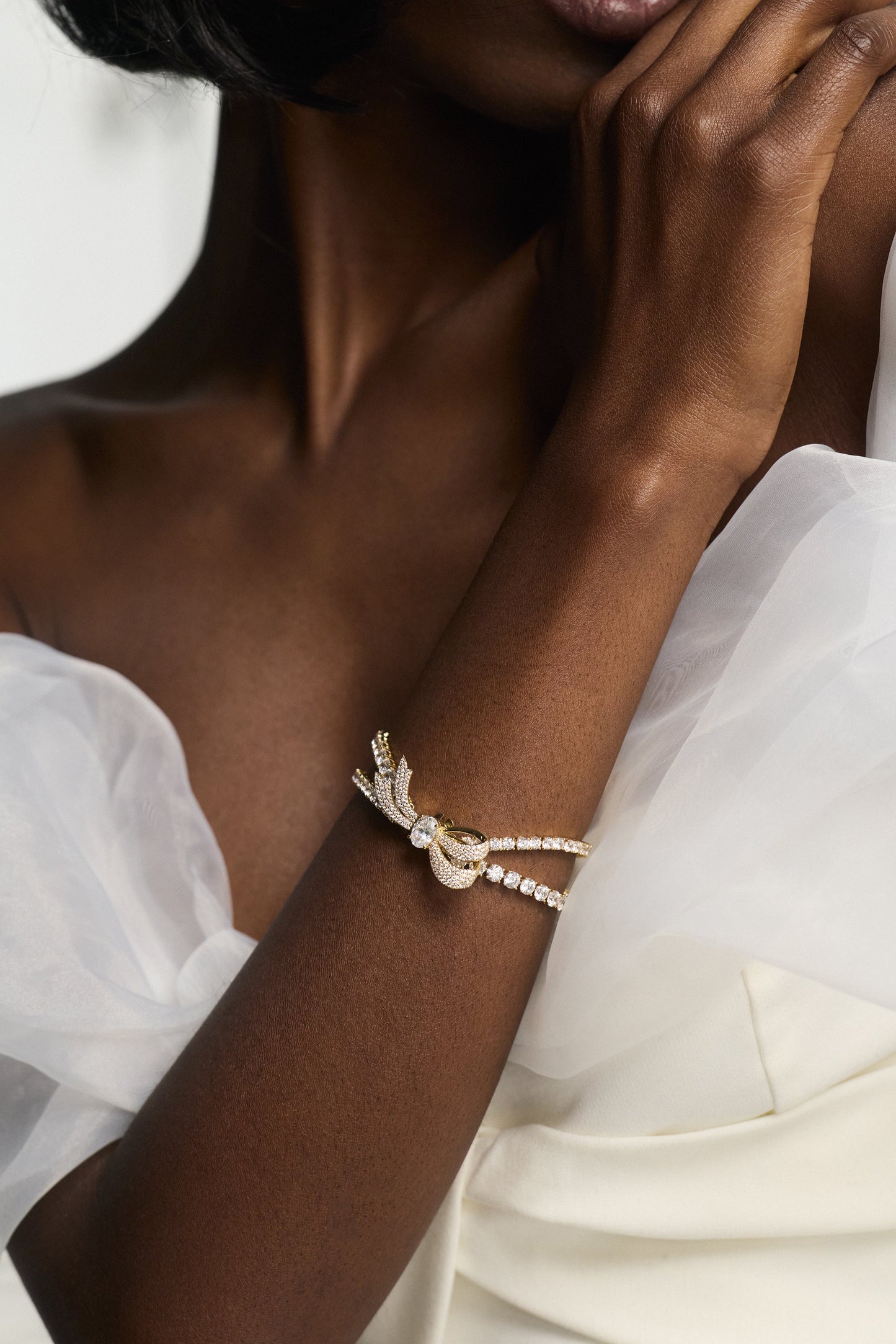 A CLOSE UP OF A MODEL’S HAND ON HER OPPOSITE SHOULDER AS SHE WEARS A WHITE DRESS AND THE EROS BOW TENNIS BRACELET