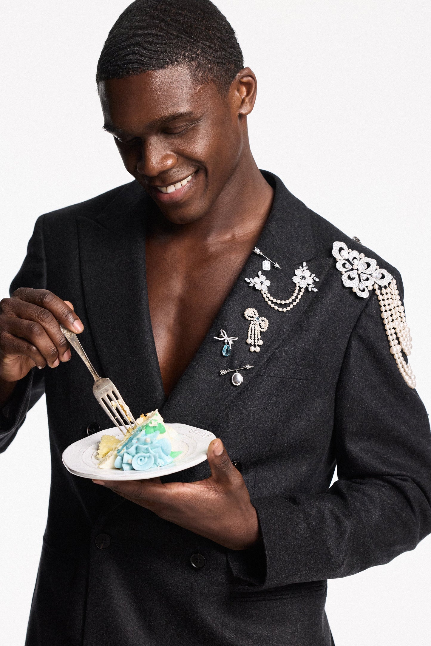  A MODEL IN A SUIT JACKET EATING A PIECE OF CAKE, WITH CHARMS PINNED TO HIS LAPEL