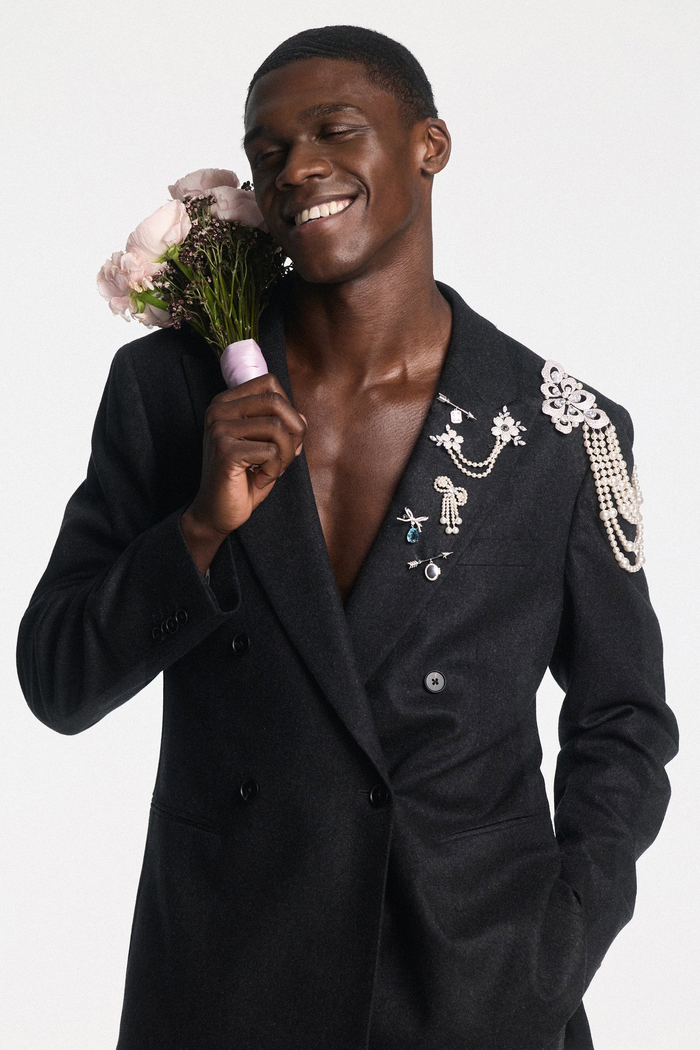 A MODEL SMILING IN A SUIT JACKET WITH CHARMS PINNED TO HIS LAPEL, WHILE HOLDING A PINK BOUQUET UP TO HIS SHOULDER