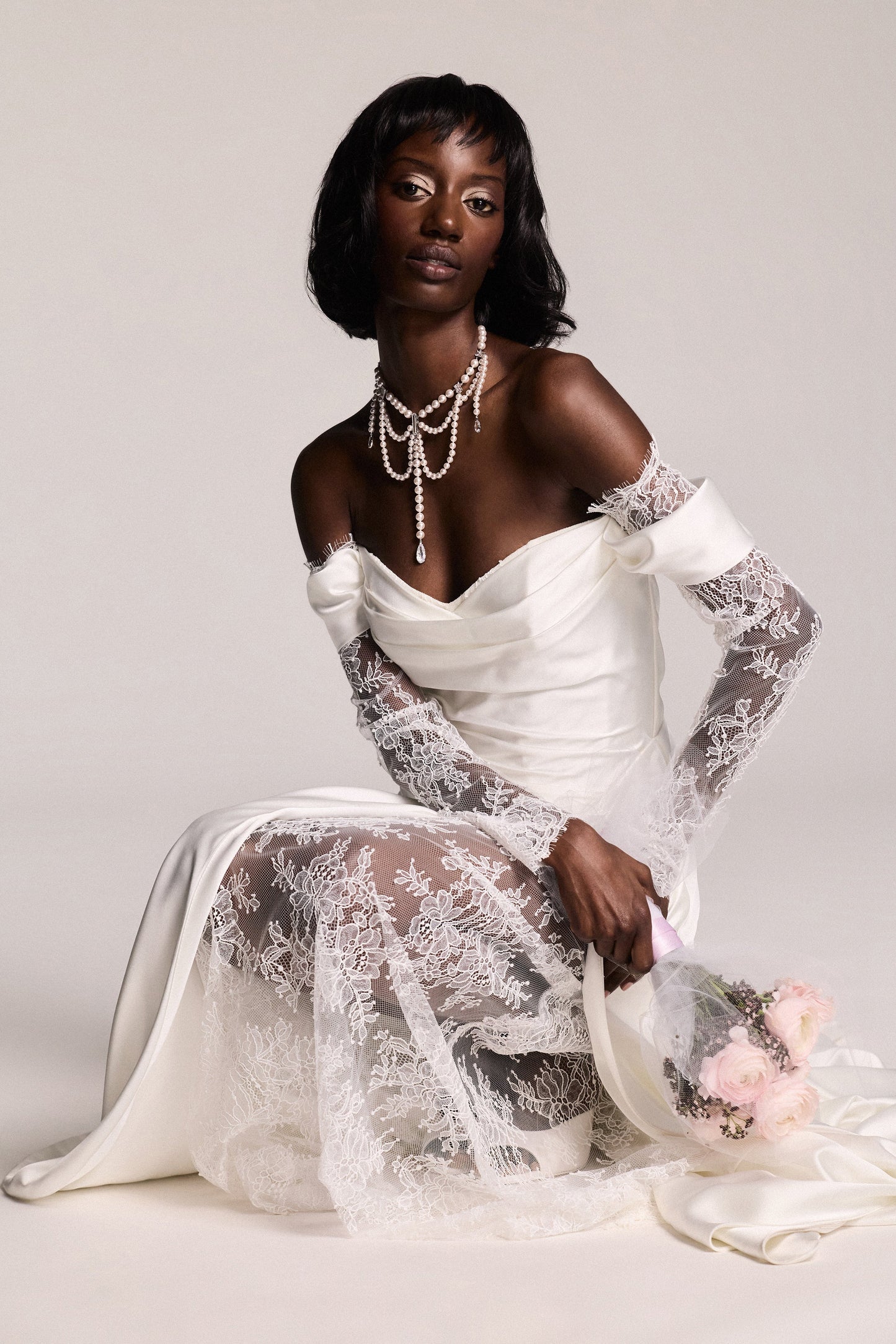 A MODEL CROUCHED DOWN IN A WEDDING DRESS AND HOLDING A PINK BOUQUET, WEARING THE PEARL CHOKER LOW DROPS NECKLACE