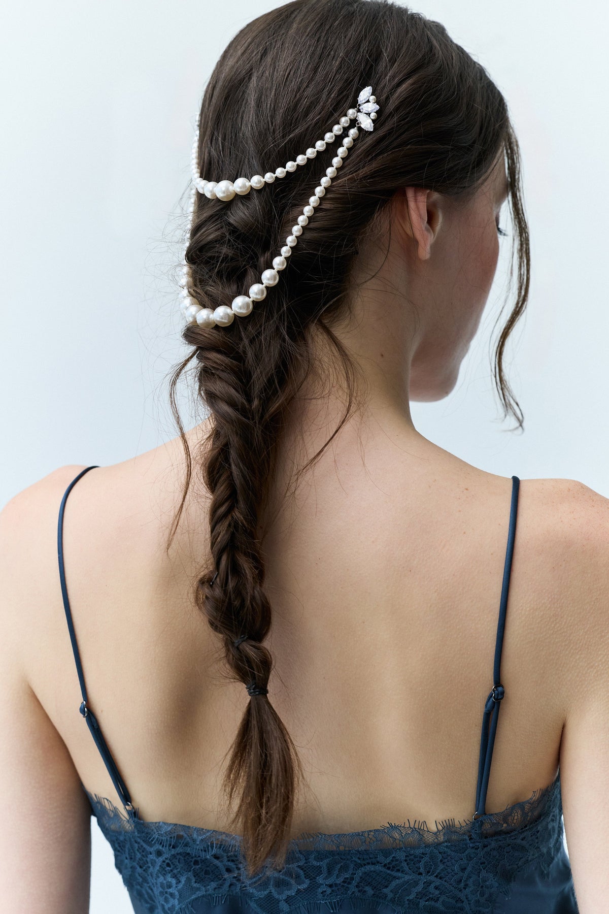 WOMAN IN A BLUE DRESS VIEWED FROM BEHIND, SLIGHTLY LOOKING DOWN WITH A LONG BRUNETTE BRAID BEHIND HER, FEATURING NADRI’S PEARL SWAG HAIR PIN PINNED ATOP HER HEAD