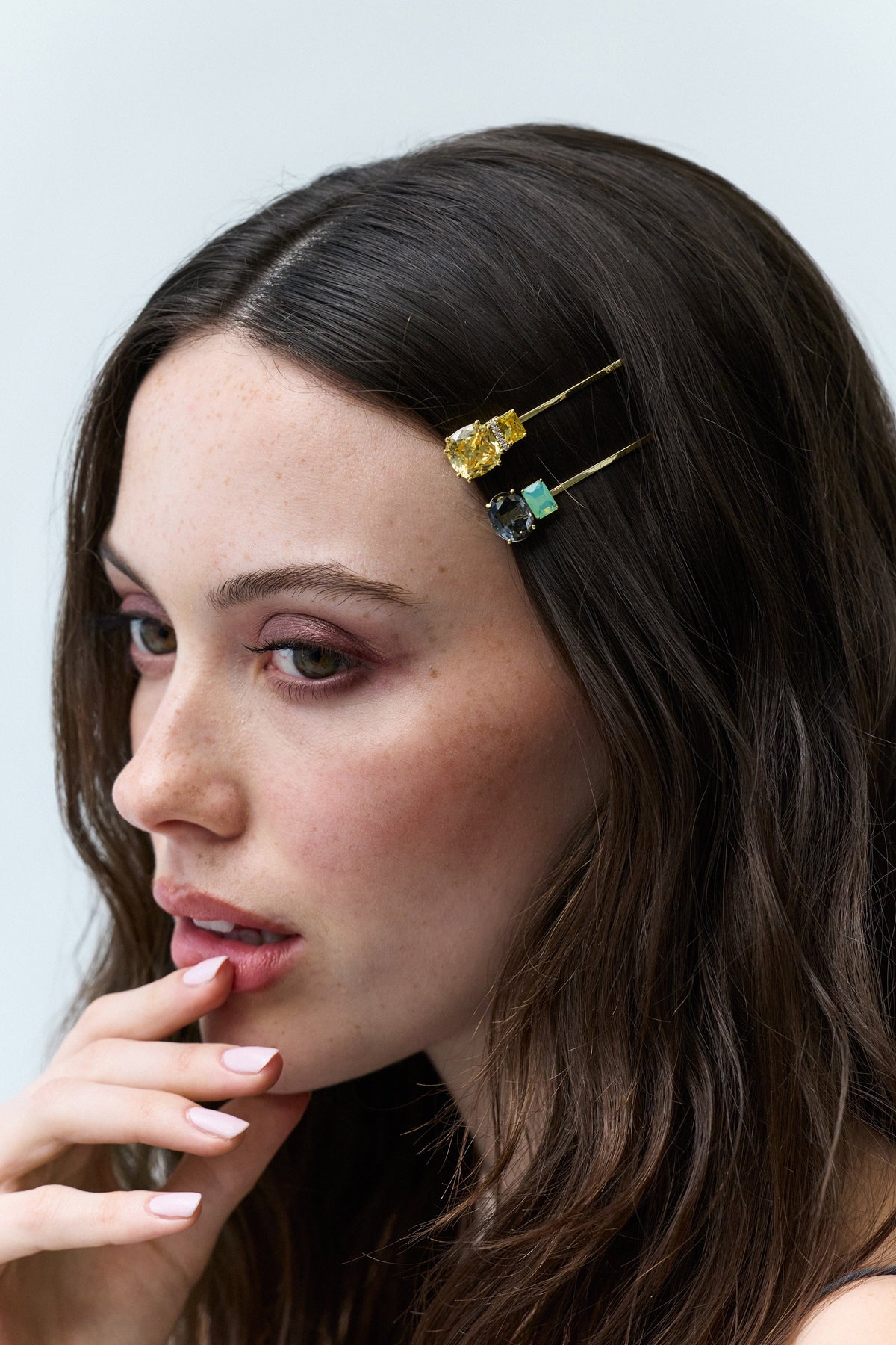 A WOMAN WITH BRUNETTE HAIR TURNED TO THE SIDE, FEATURING A YELLOW AND GREEN BOBBY PIN FROM NADRI'S HAIR TODAY COLLECTION PINNING HER HAIR BACK, AS HER HAND RESTS GENTLY AGAINST HER LIPS.