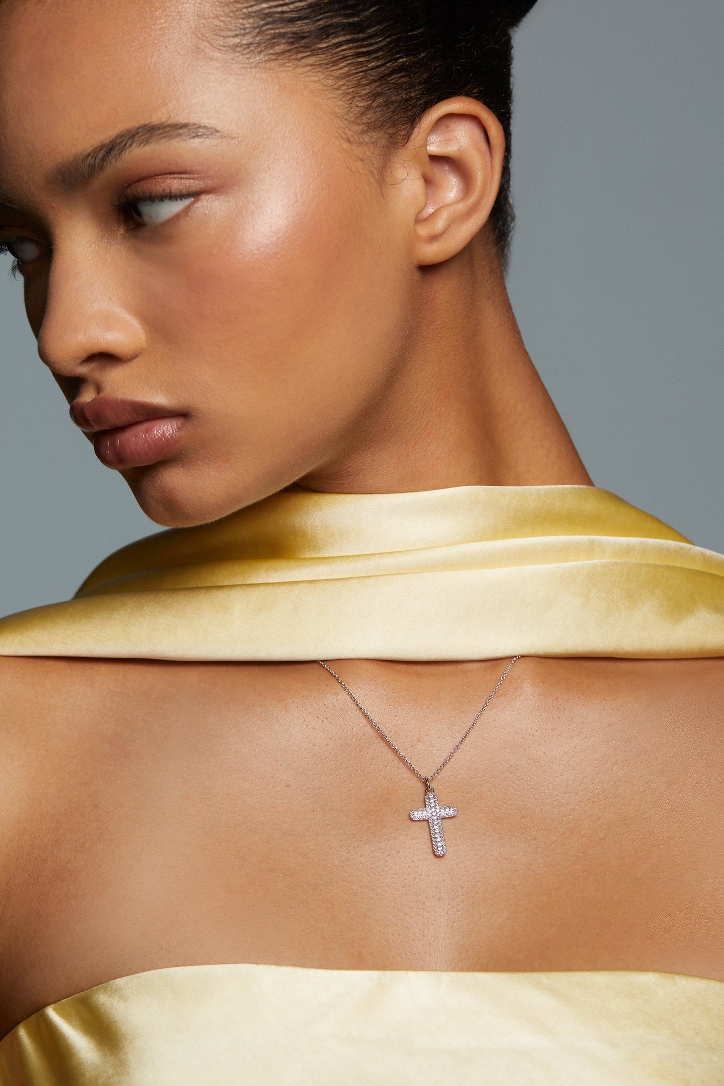 CLOSE UP OF MODEL WEARING CRUSHED CROSS PENDANT NECKLACE. SHE HAS ON A STRAPLESS SILK YELLOW DRESS AND MATCHING SCARF.  HER HAIR IS UP IN A TIGHT BUN AND SHE IS FACING AWAY FROM THE CAMERA.