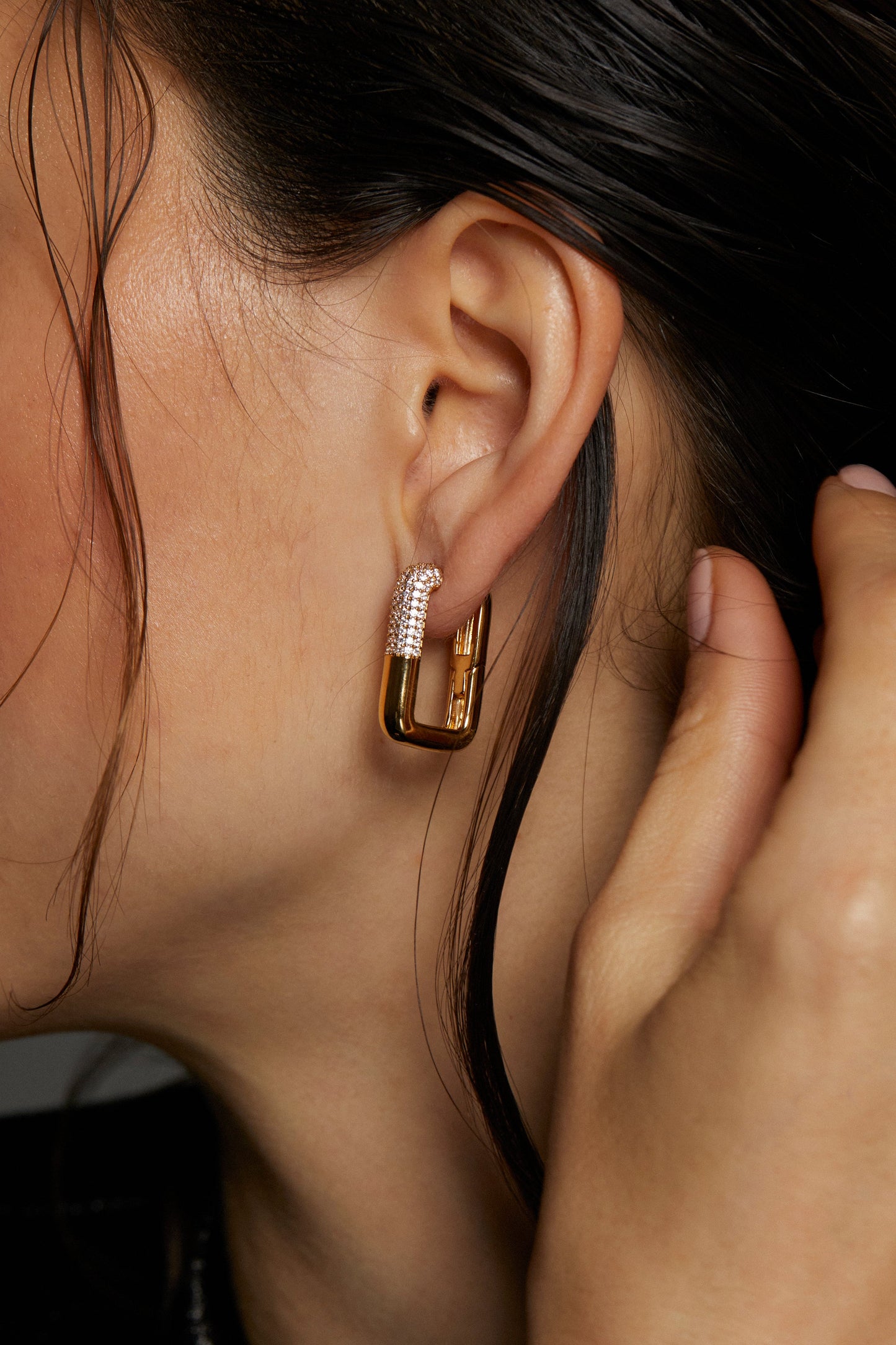 CLOSE UP IMAGE OF ALMA SQUARE HOOP EARRINGS ON MODEL. HER FACE IS CROPPED OUT AND HER HAIR IS TUCKED BEHIND HER EAR. HER HAND IS REACHING UP TO HER EAR TO PULL HER HAIR BACK.