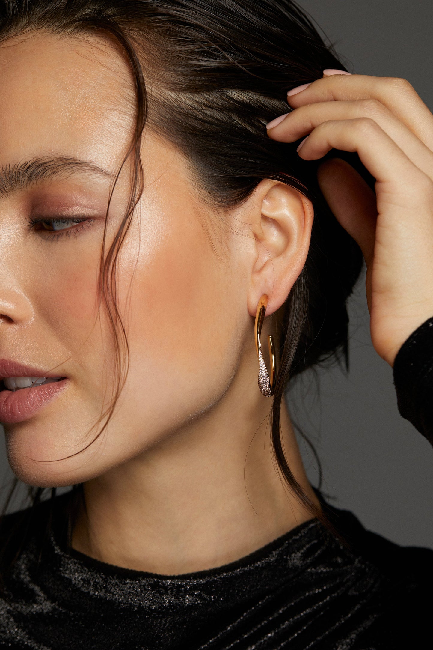MODEL WEARING ALMA LARGE RIBBON C HOOP EARRINGS. IT IS A CLOSE OF IMAGE OF HALF HER FACE. HER HAND IS PULLING HER HAIR BACK BEHIND HER EARS AND HER EYE IS HALF CLOSED. 