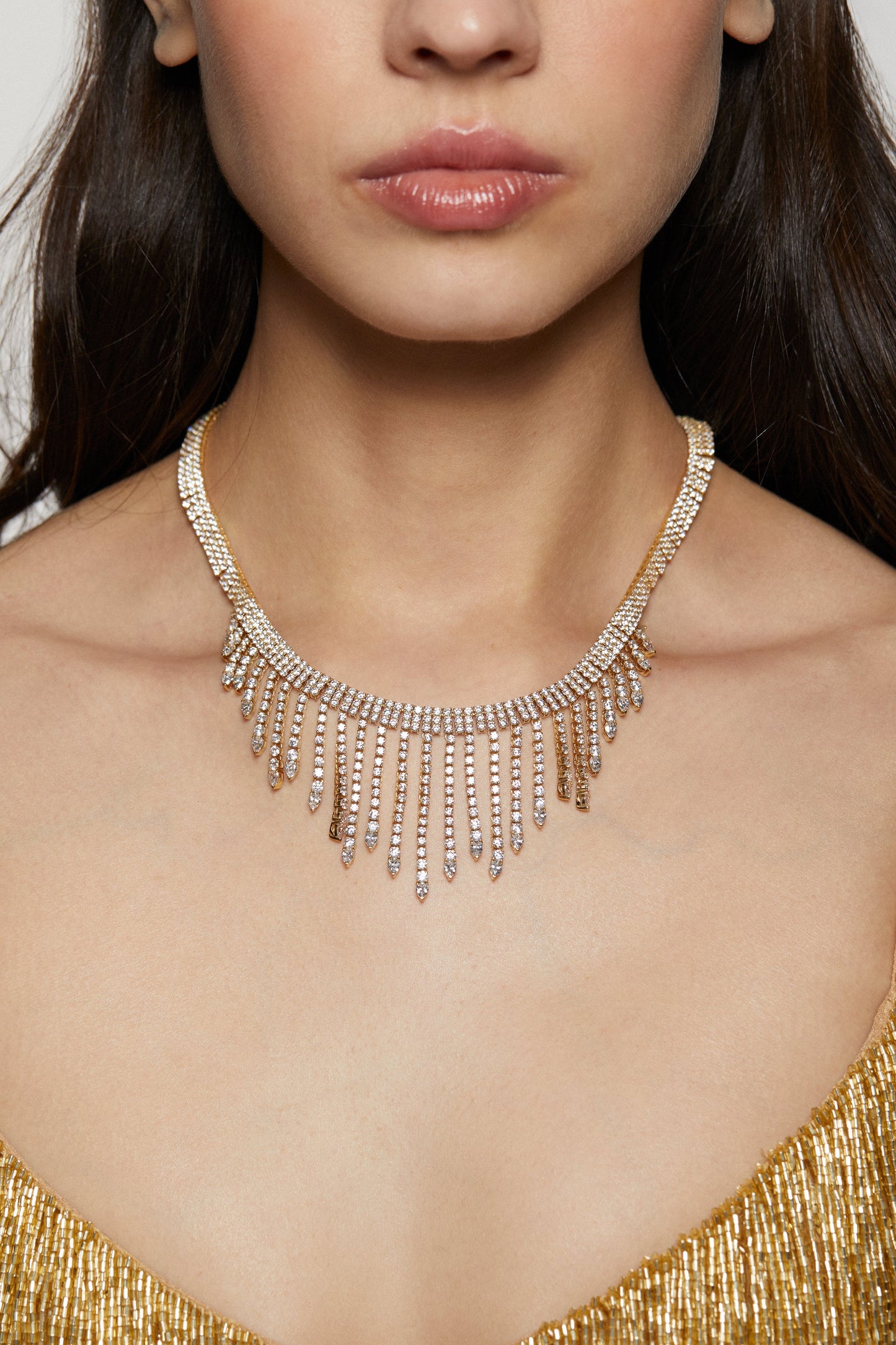 MODEL WEARING DIVA FRINGE COLLAR NECKLACE. IT IS A CLOSE UP IMAGE OF HER NECKLINE. HER HAIR IS BEHIND HER SHOULDERS. SHE IS WEARING A BEADED GOLD SHIMMERING DRESS. 