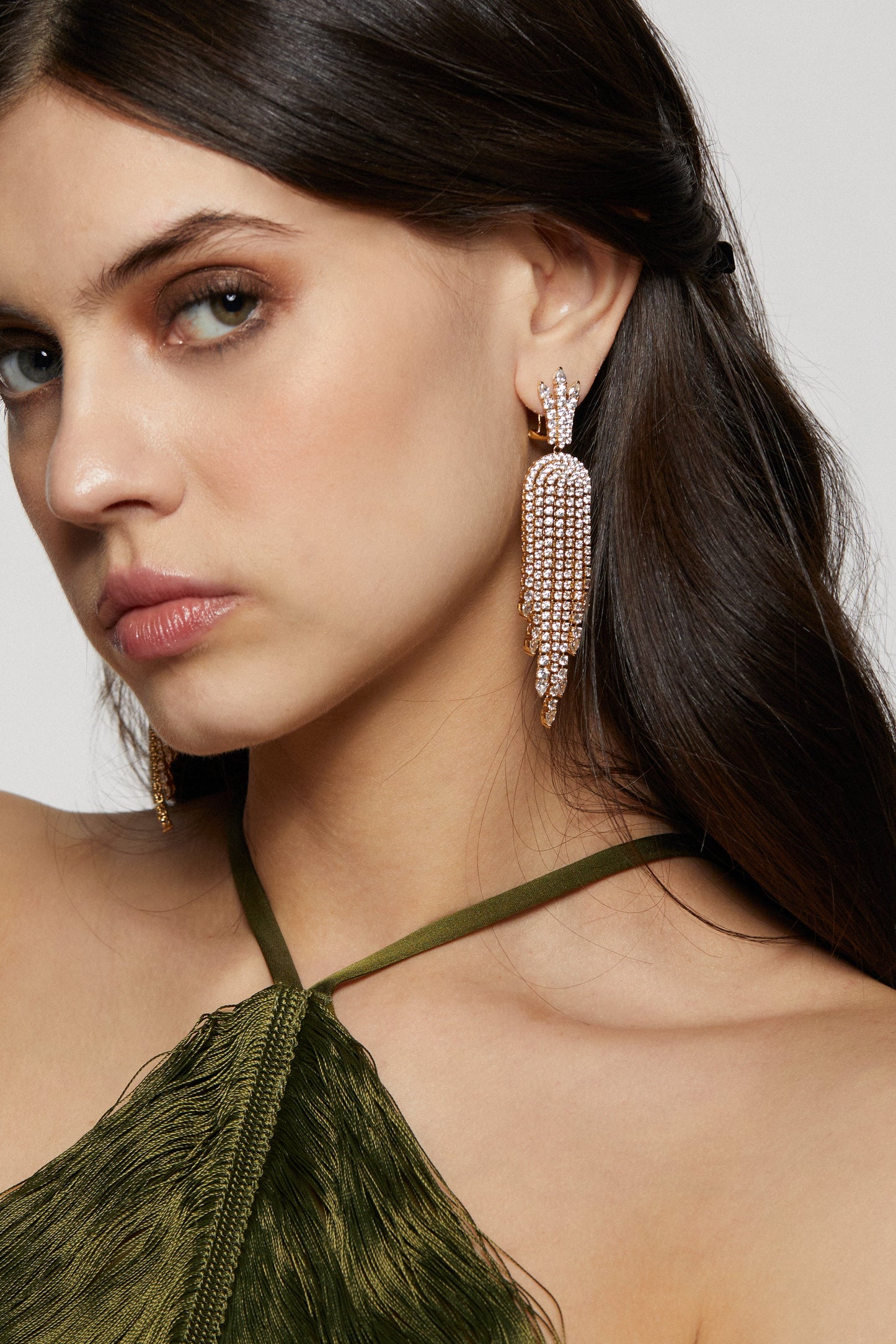 DIVA FRINGE CHANDELIER EARRINGS ON MODEL. SHE IS LOOKING TOWARDS THE CAMERA WITH HER HAIR PINNED BACK AND OFF THE SHOULDER. SHE IS WEARING A GREEN FRINGE TOP. 