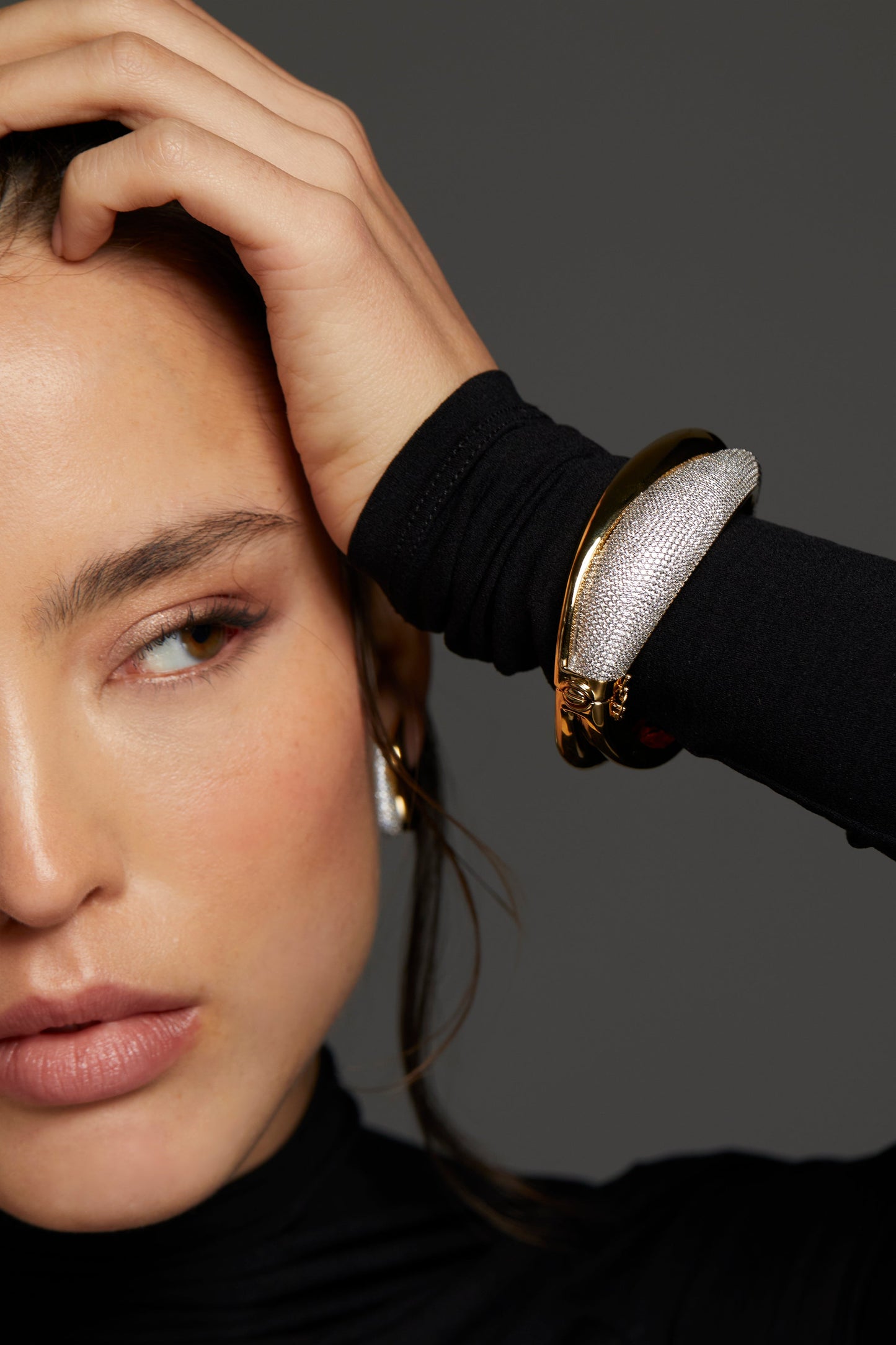 MODEL WEARING DRIP SIDE HINGE BRACELET. IT IS A CLOSE UP IMAGE OF HALF HER FACE WITH HER HAND REACHING UP TO HER HEAD SHOWING A CLOSE UP VIEW OF THE BRACELET. SHE IS WEARING A BLACK LONG SLEEVE TURTLENECK TOP. 