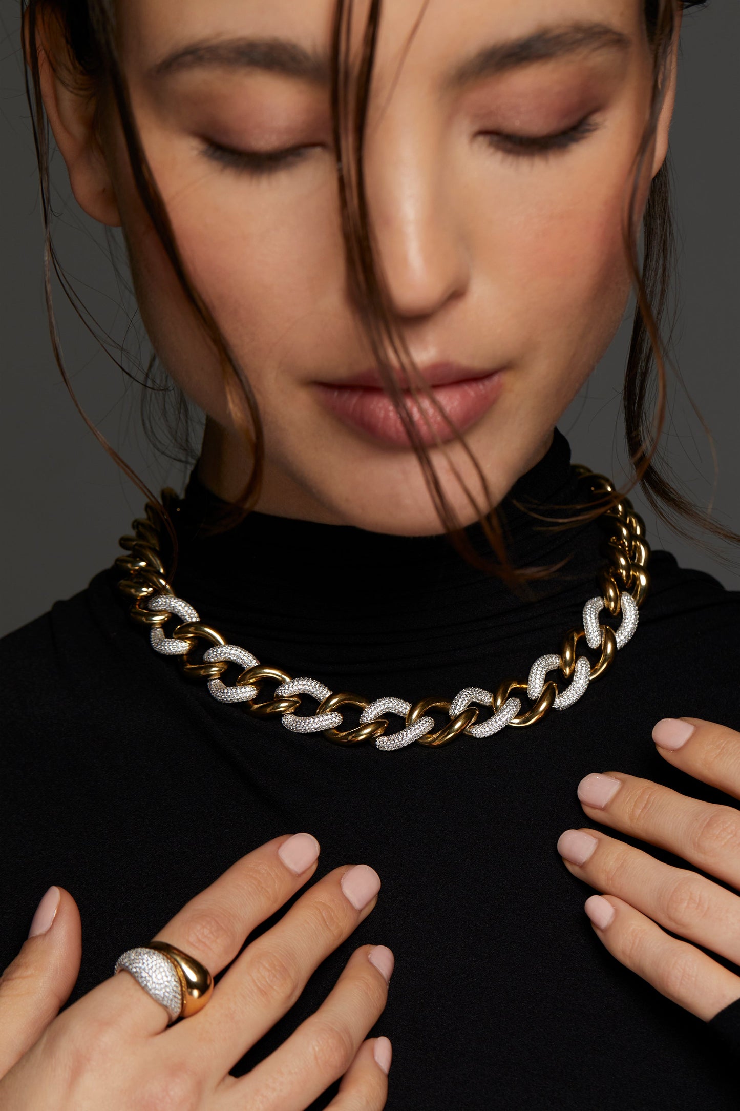 MODEL WEARING DRIP LARGE CURB CHAIN NECKLACE. HER EYES ARE CLOSED, HAIR IS PULLED BACK IN A BUN WITH LOOSE STRANDS AROUND HER FACE, EYES CLOSED, AND HANDS REACHING TOWARDS THE NECKLACE. SHE IS WEARING A BLACK TURTLENECK TOP. 