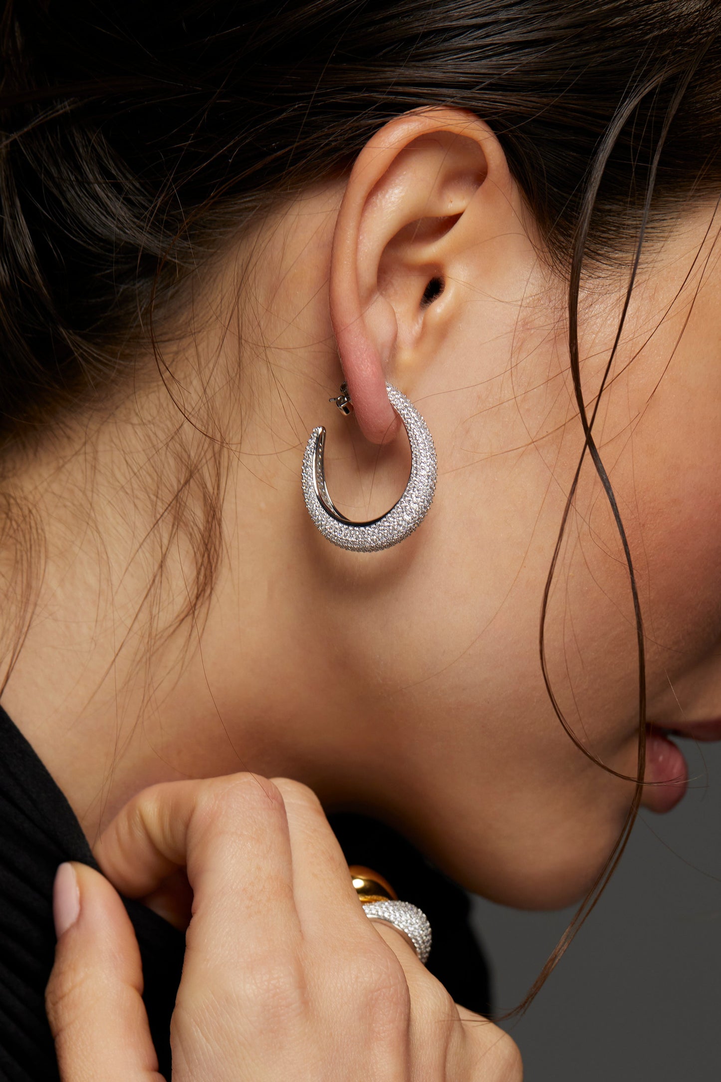 CLOSE UP  IMAGE OF MODEL WEARING DRIP PUFFY PAVE C HOOP EARRINGS. SHE IS FACING TO THE SIDE AND IT IS A TIGHT SHOT OF HER EAR SHOWING A SIDE VIEW OF THE HOOP. HER HAIR IS IN A LOOSE BUN AND HER HAND IS REACHING TOWARDS HER BLACK TURTLENECK TOP.