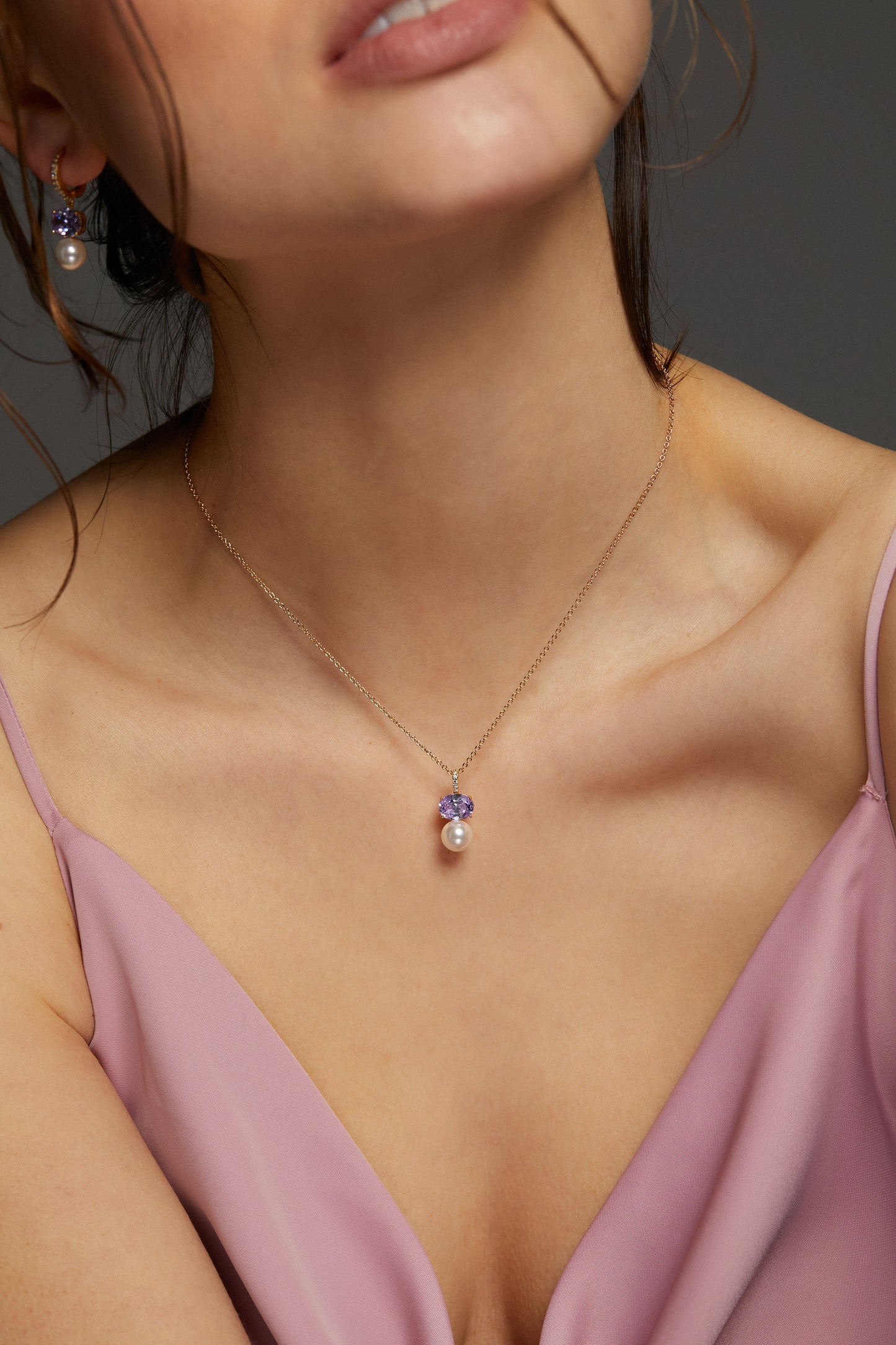 MODEL WEARING NATURAL BEAUTY LAVENDER PEARL PENDANT. SHE HAS ON A PINK V-NECK DRESS. IT IS A CLOSE UP IMAGE OF HER NECKLINE WITH HER EAR IN SHOT OF THE MATCHING EARRINGS TO THE NECKLACE. 